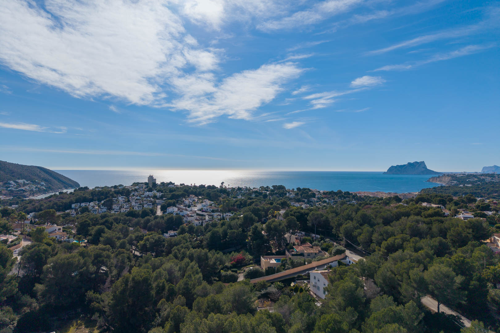 Vue aérienne panoramique de la côte de Moraira, révélant une pinède dense parsemée de villas et la silhouette du Peñón de Ifach sur l'horizon méditerranéen.