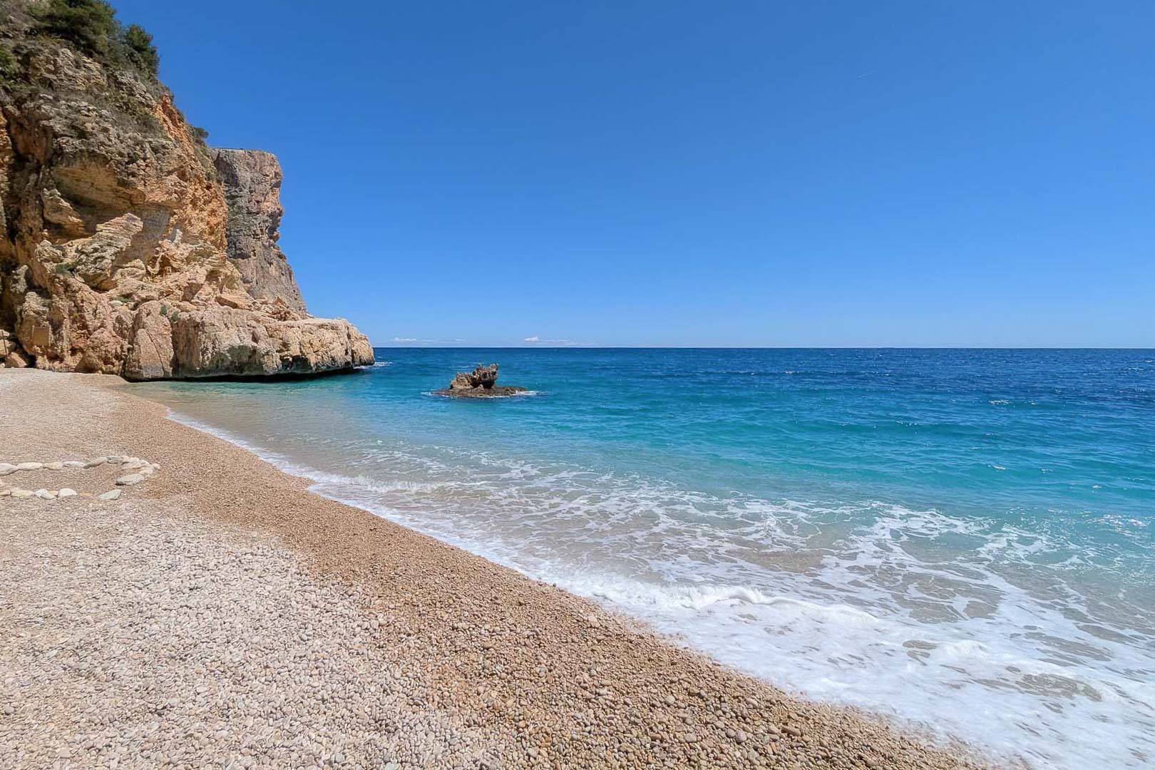 Vue d'une crique aux eaux cristallines et plage de galets, bordée de falaises calcaires sous un ciel azur près de Moraira.