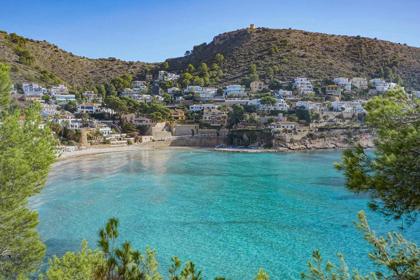 Vue panoramique sur la baie d'El Portet à Moraira, avec ses eaux turquoise, sa plage de sable et ses villas méditerranéennes nichées sur la colline sous la tour de guet.