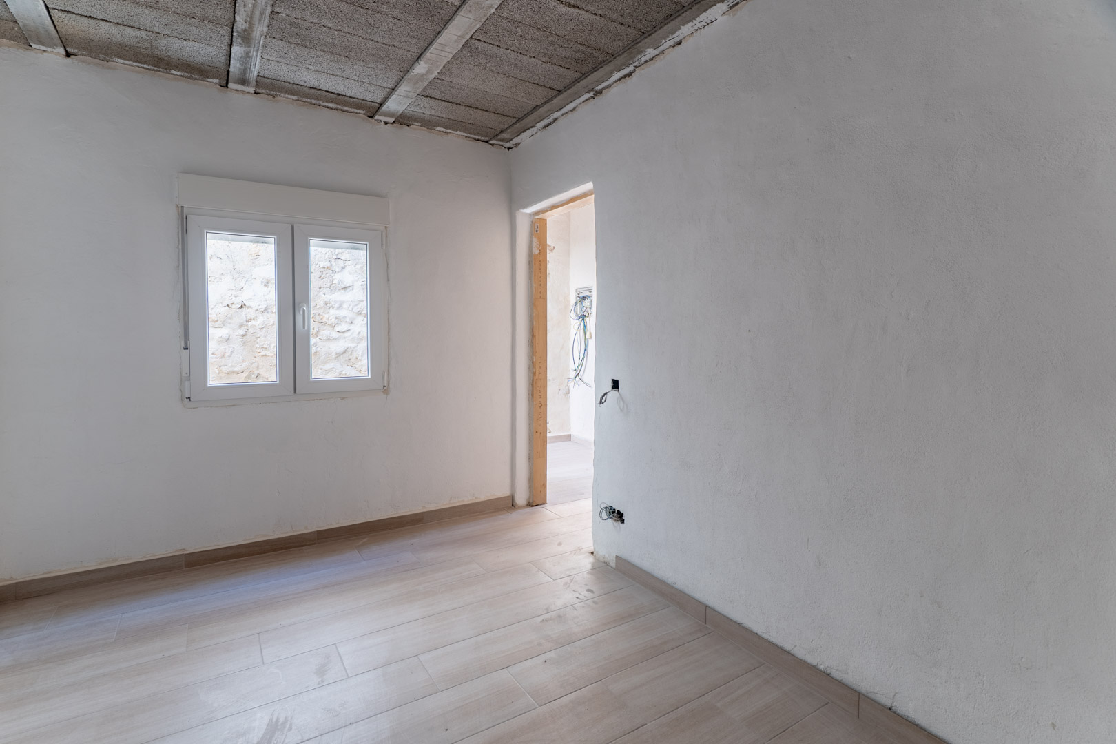 Espace intérieur en rénovation avec sol en grès cérame imitation bois, murs blancs et fenêtre PVC. Le plafond révèle la structure porteuse en béton.