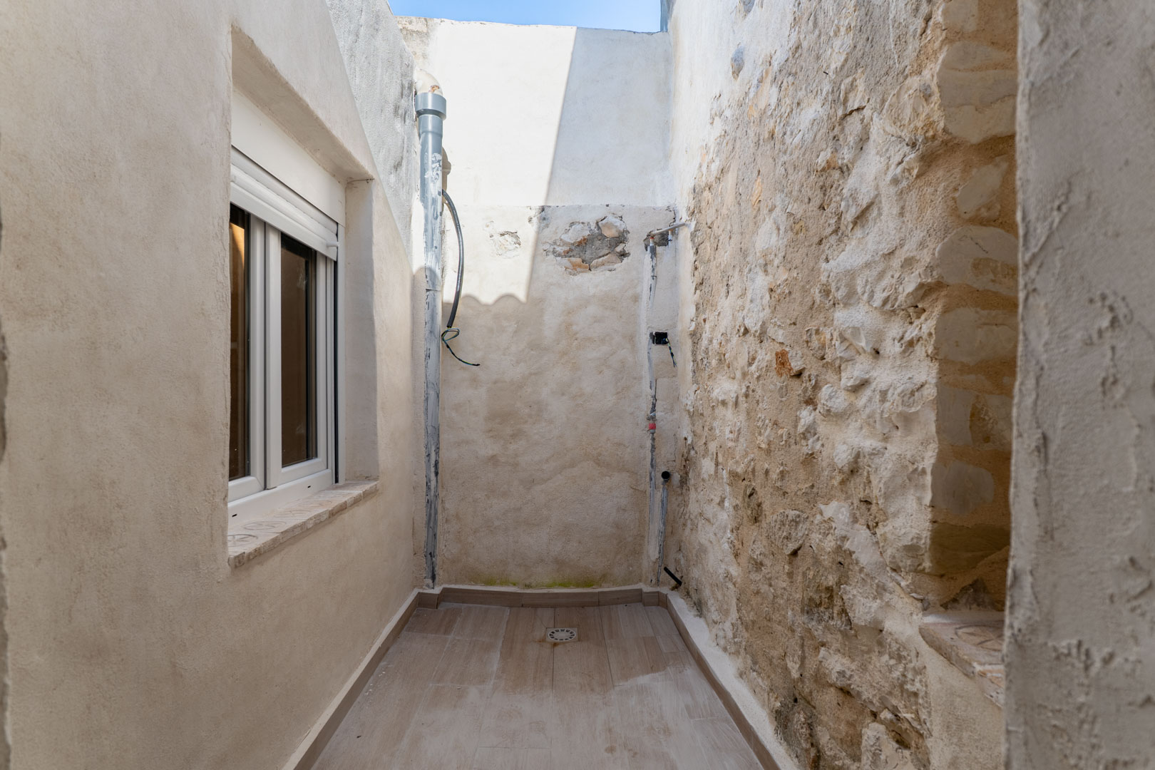 Patio intérieur étroit avec sol en céramique et évacuation, alliant un mur d'origine en pierre sèche à une façade rénovée avec fenêtre en PVC blanc.