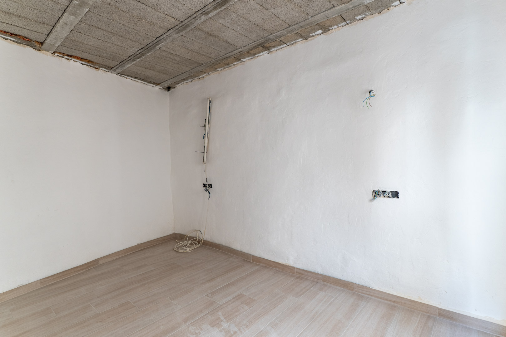 Pièce en cours de rénovation avec sol en grès cérame imitation bois, murs blancs et plafond en poutrelles de béton avec pré-installation électrique.