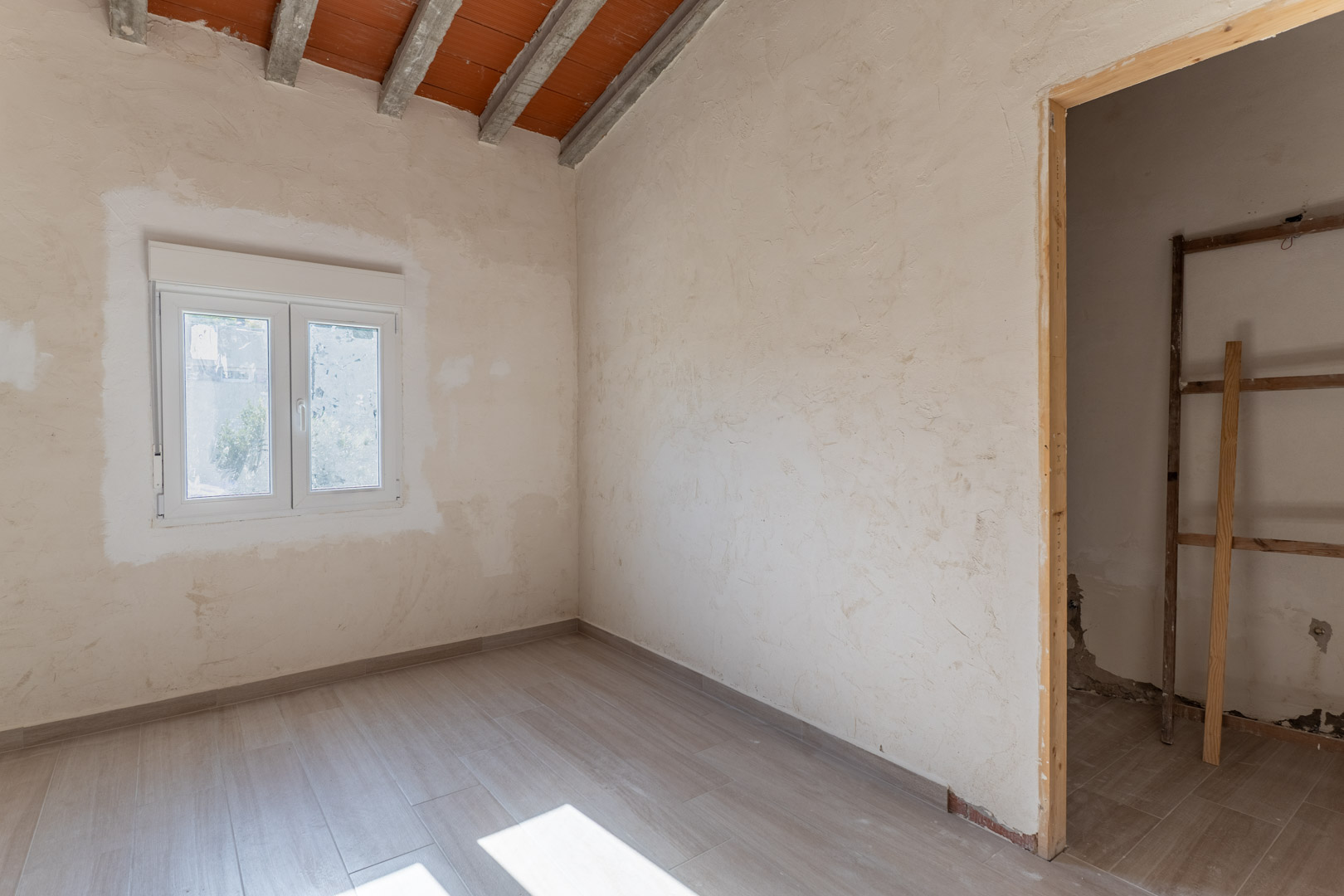 Pièce en rénovation avec sol en grès cérame aspect bois, fenêtre en PVC blanc et plafond incliné aux poutres en béton et briques apparentes.