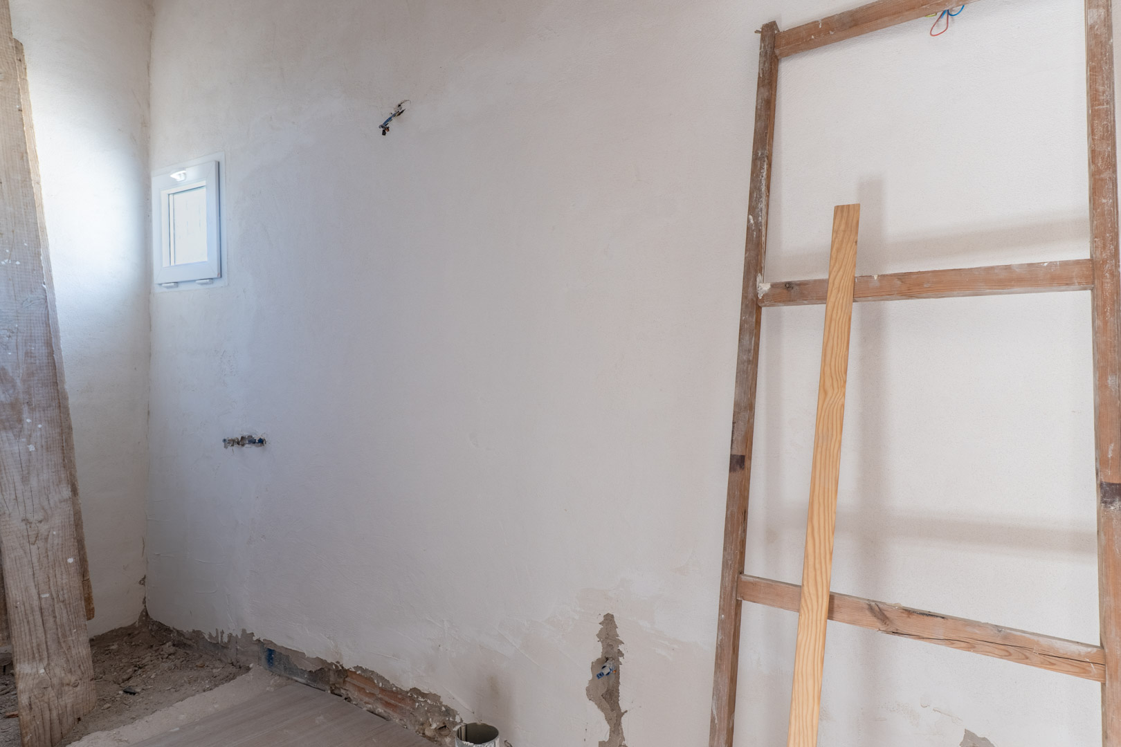 Pièce intérieure en cours de rénovation avec murs plâtrés, petite fenêtre blanche et raccordements électriques en attente de finition.