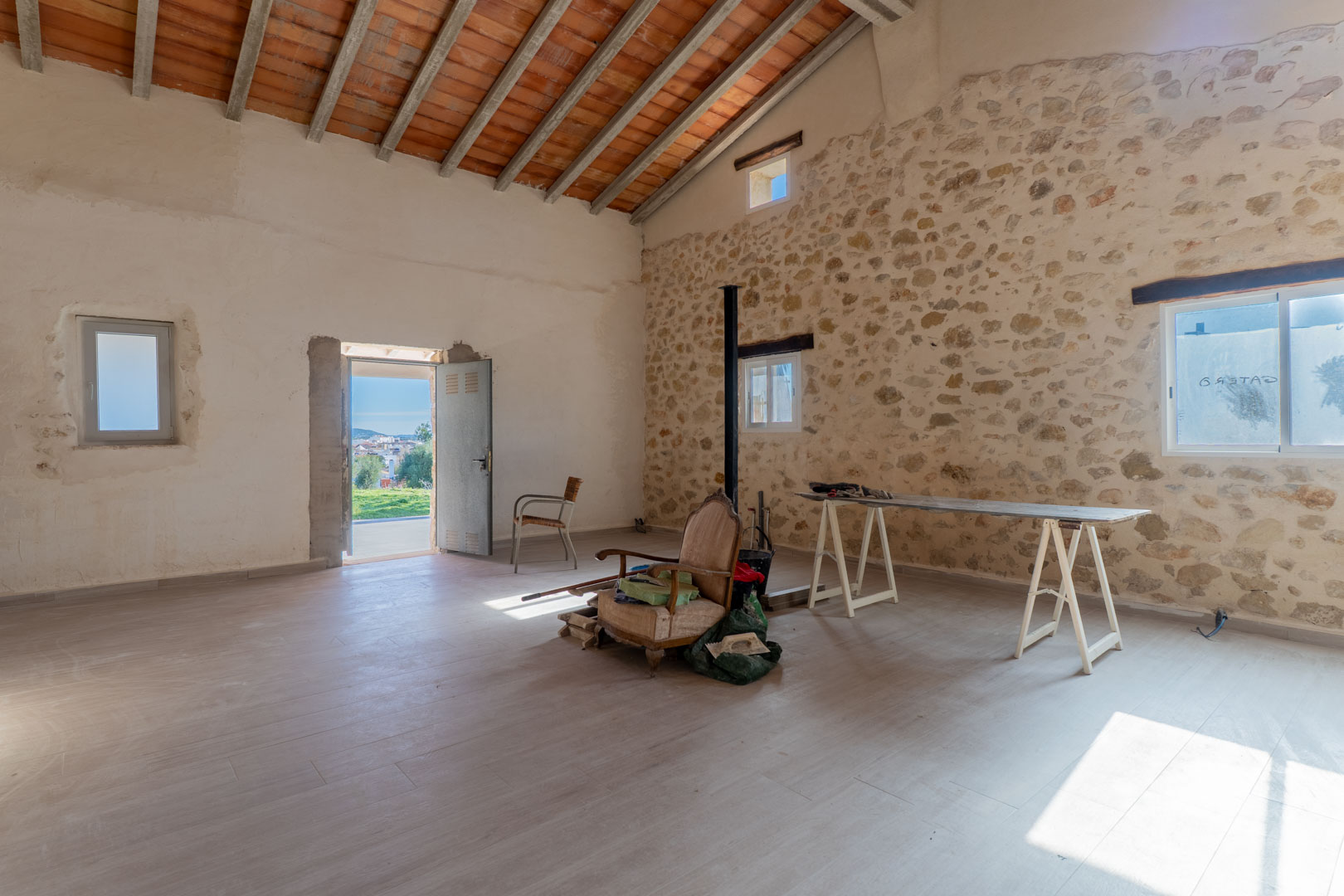 Pièce principale spacieuse avec murs en pierre sèche, hauts plafonds à poutres apparentes et accès direct vers l'extérieur offrant une luminosité naturelle.