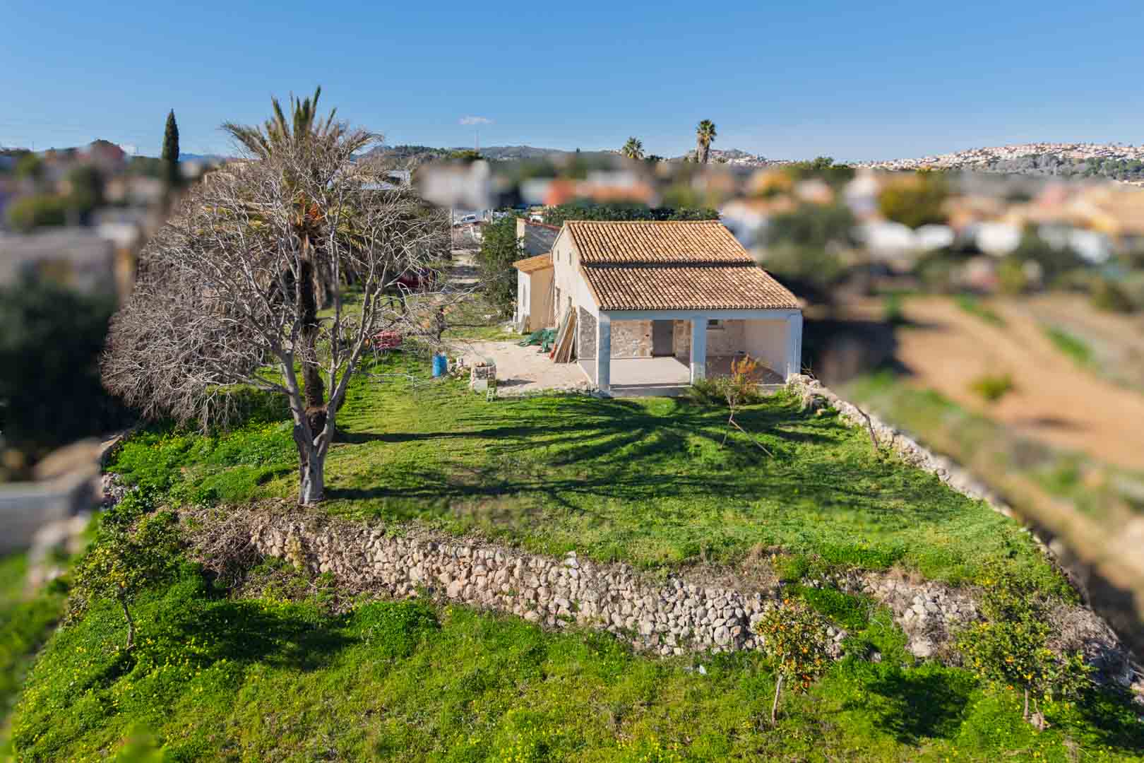 Vue aérienne de la finca traditionnelle avec son toit rénové, ses murs en pierre sèche et sa naya spacieuse, entourée d'un jardin verdoyant avec des arbres fruitiers.