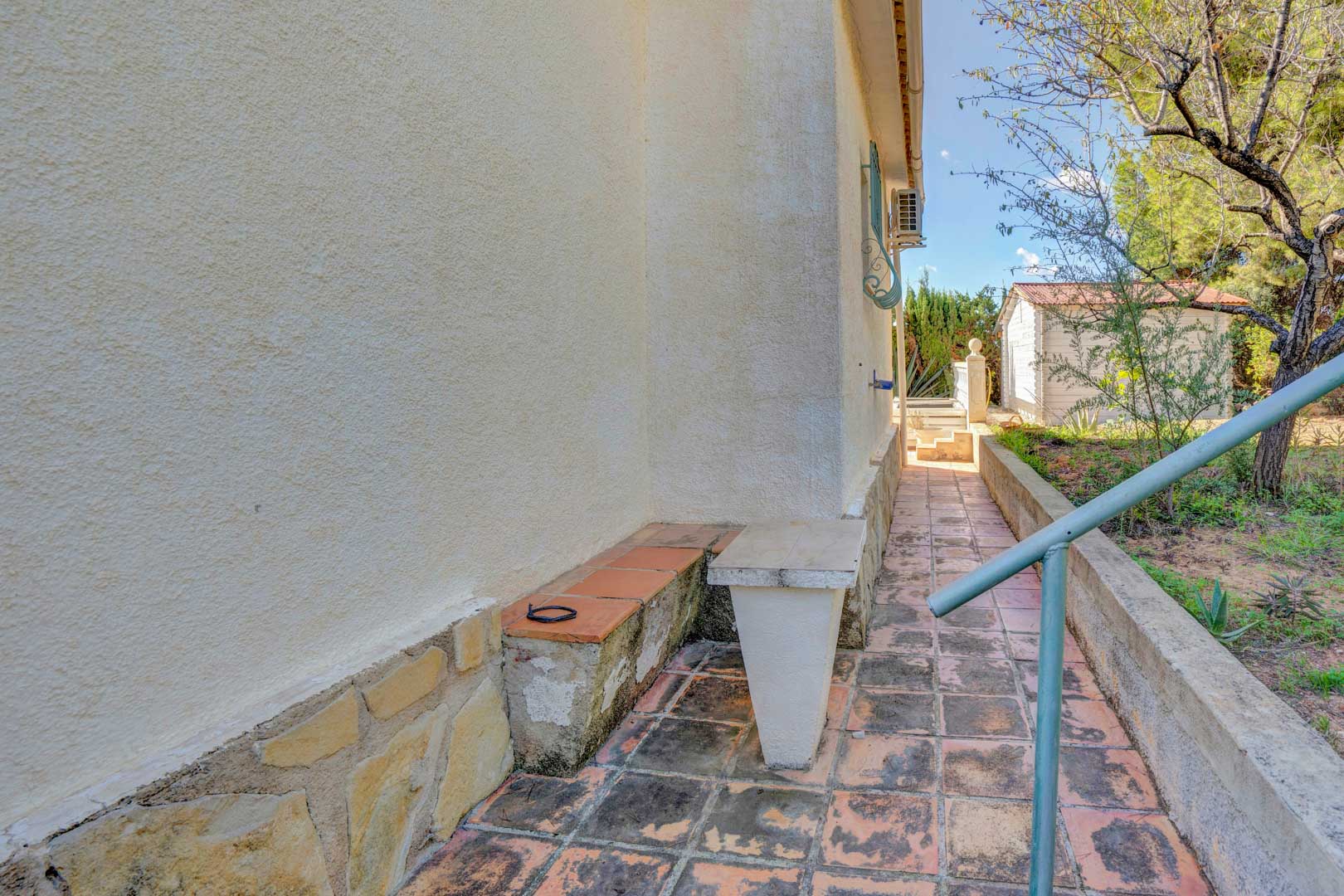 Allée latérale extérieure avec revêtement en terre cuite, soubassement en pierre et vue sur le jardin arboré avec cabanon de rangement.