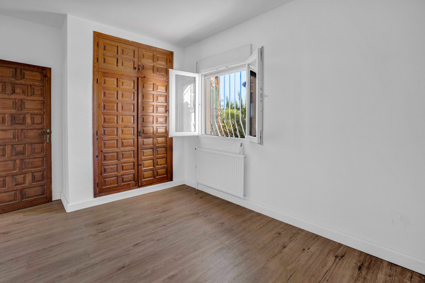 Chambre lumineuse avec placard intégré en bois sculpté, sol stratifié, radiateur et fenêtre en PVC offrant une vue sur la végétation extérieure.