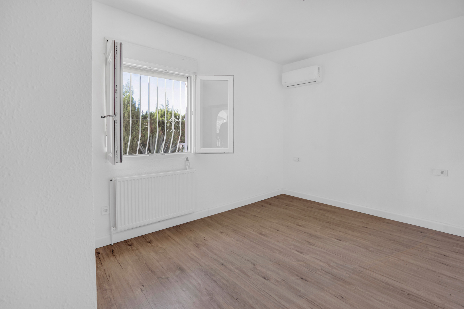 Chambre lumineuse avec sol en parquet stratifié, équipée d'un radiateur de chauffage central et d'un climatiseur split sur des murs blancs.