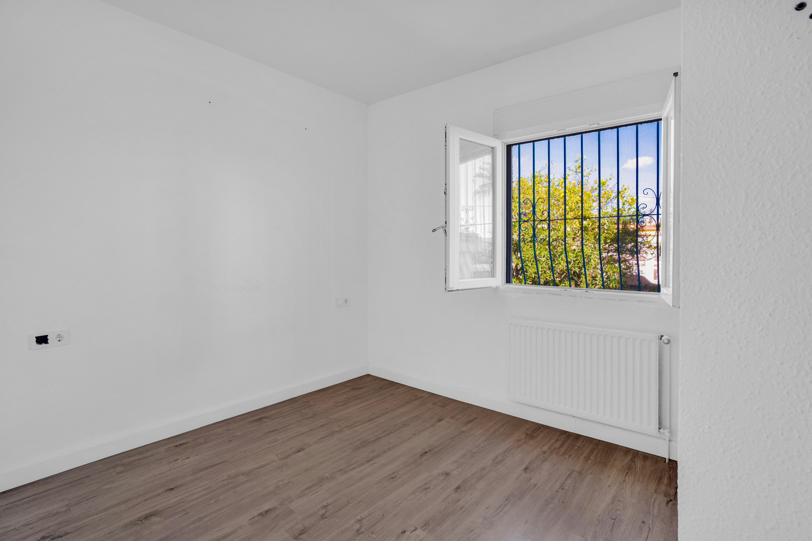 Chambre lumineuse avec sol en stratifié effet bois, murs blancs et fenêtre avec grilles décoratives offrant une vue sur la végétation et un radiateur.