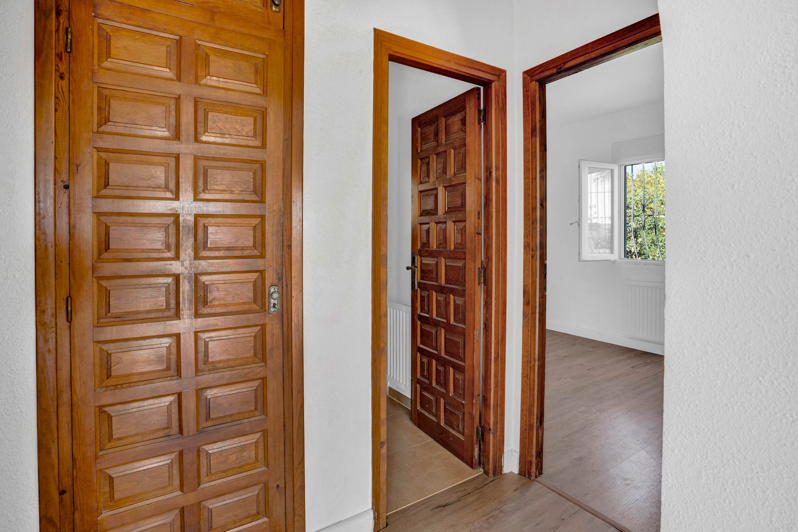 Couloir de distribution avec des portes en bois massif sculpté, des murs blancs et un sol stratifié menant aux chambres lumineuses avec vue sur le jardin.