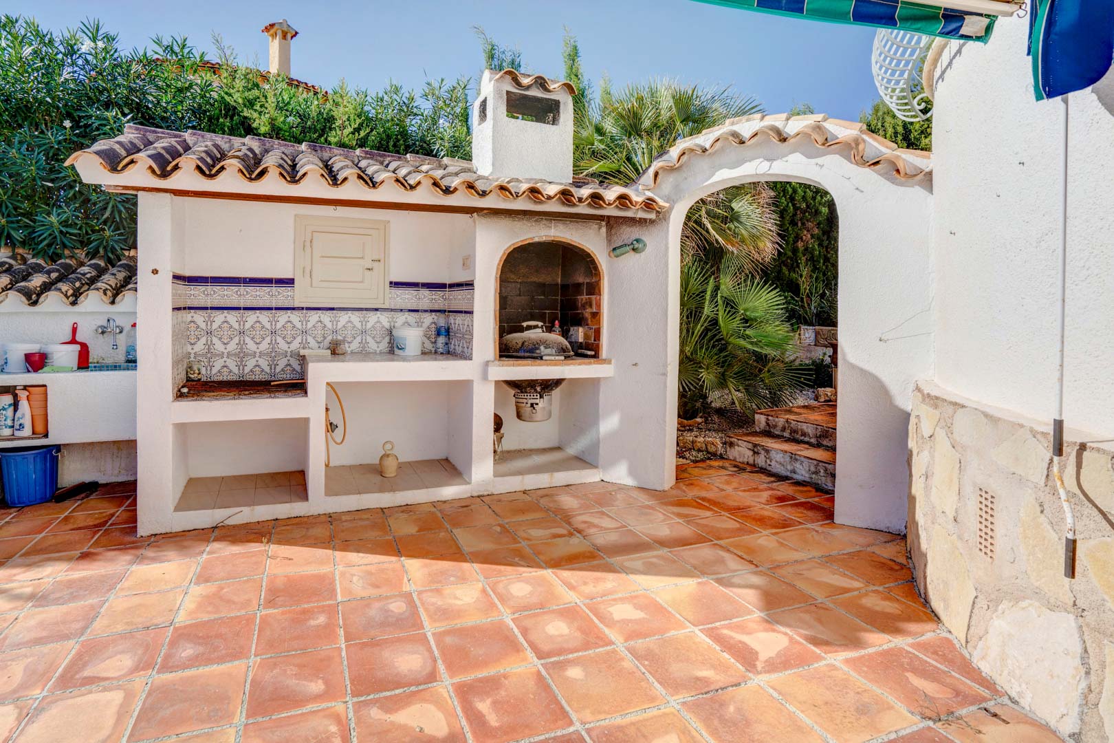 Espace barbecue maçonné avec cheminée, plan de travail en faïence traditionnelle et sol en terre cuite, situé à côté d'une arche menant au jardin.