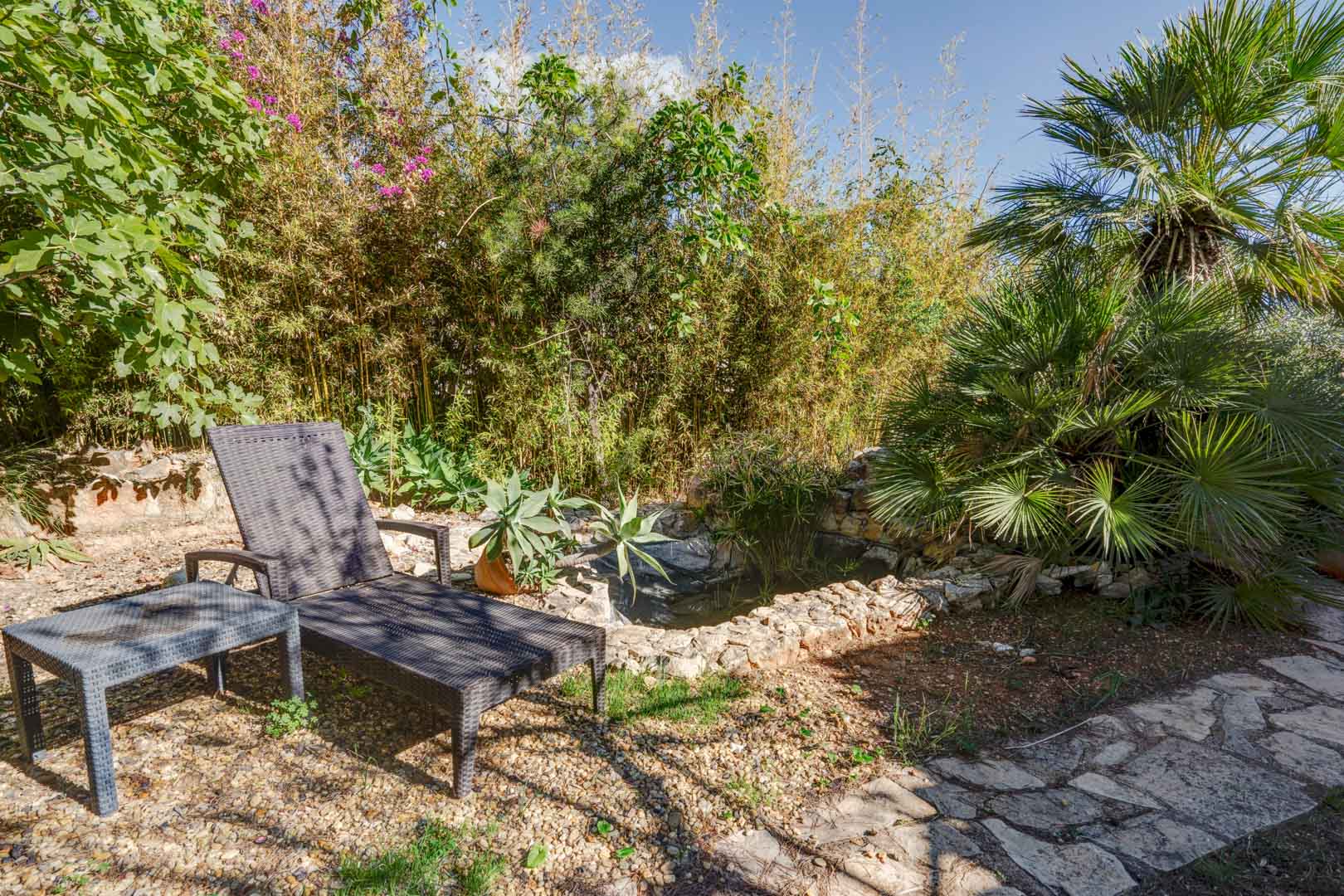 Espace de détente dans le jardin avec chaise longue en résine tressée près d'un bassin en pierre, entouré d'une végétation dense de bambous et de palmiers.
