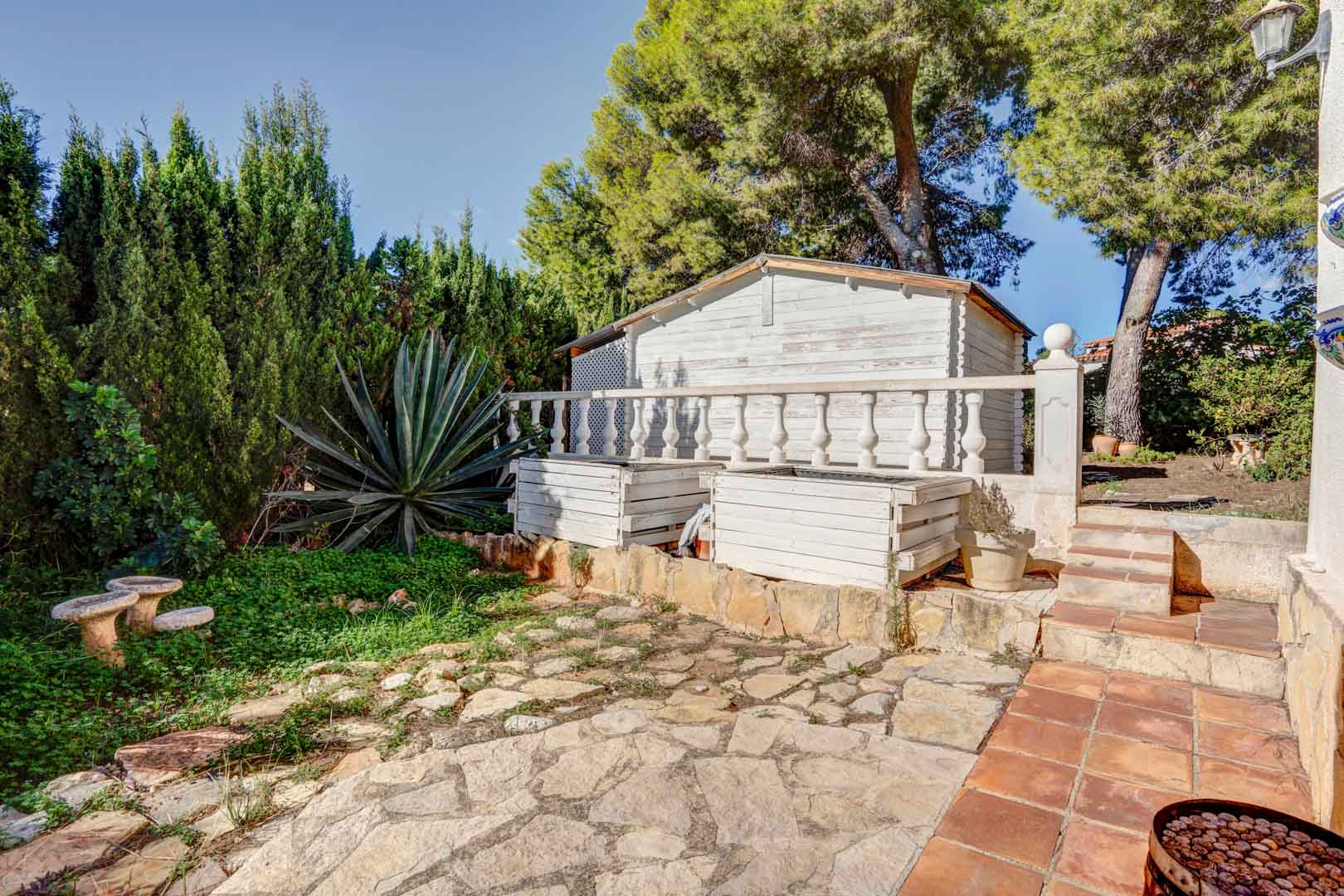 Espace extérieur avec pavage en pierre naturelle, abri de jardin en bois blanc et végétation méditerranéenne composée de pins et d'agaves.