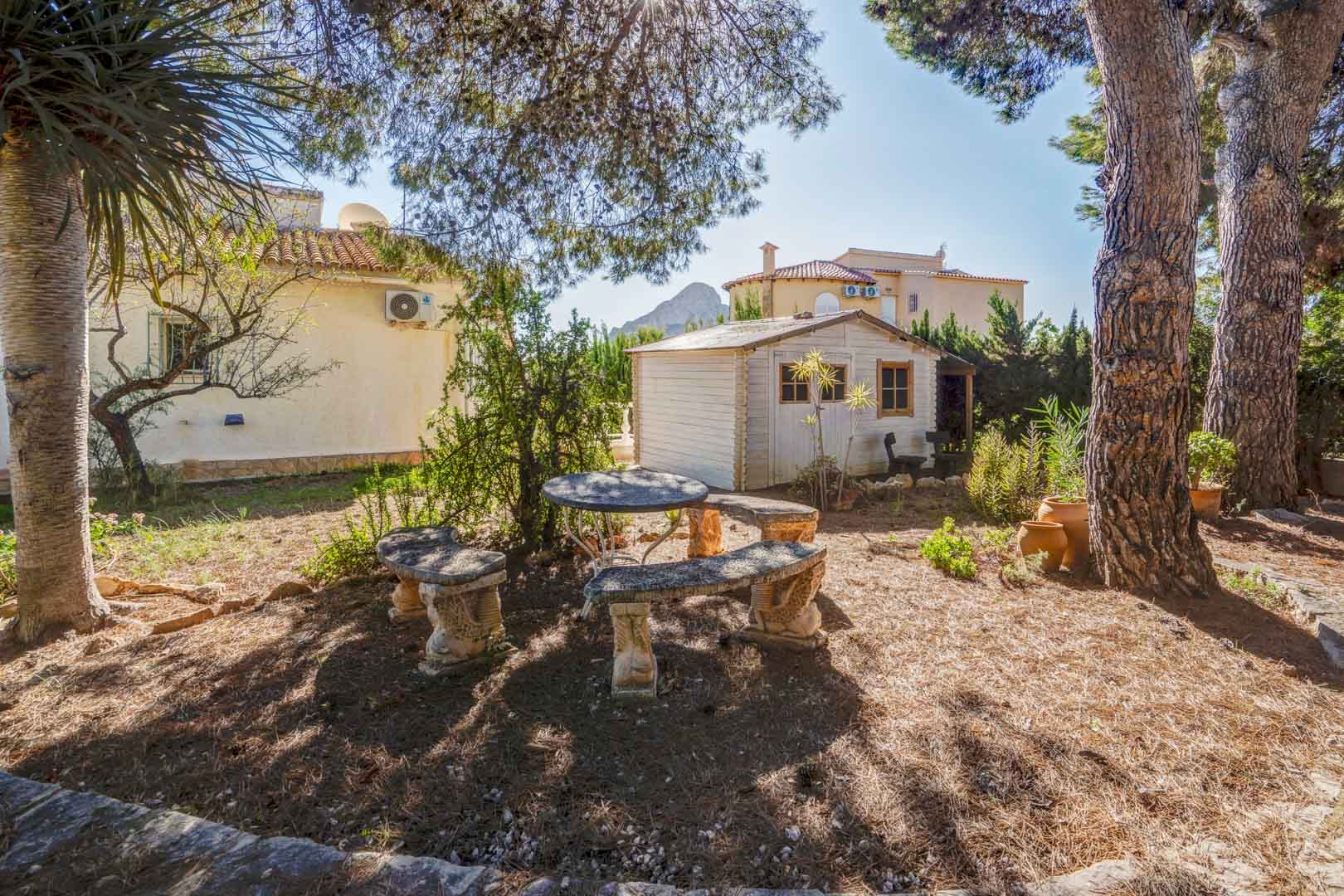 Jardin méditerranéen avec coin salon en pierre sous les pins, abri de jardin en bois blanc et vue lointaine sur le Peñón de Ifach.
