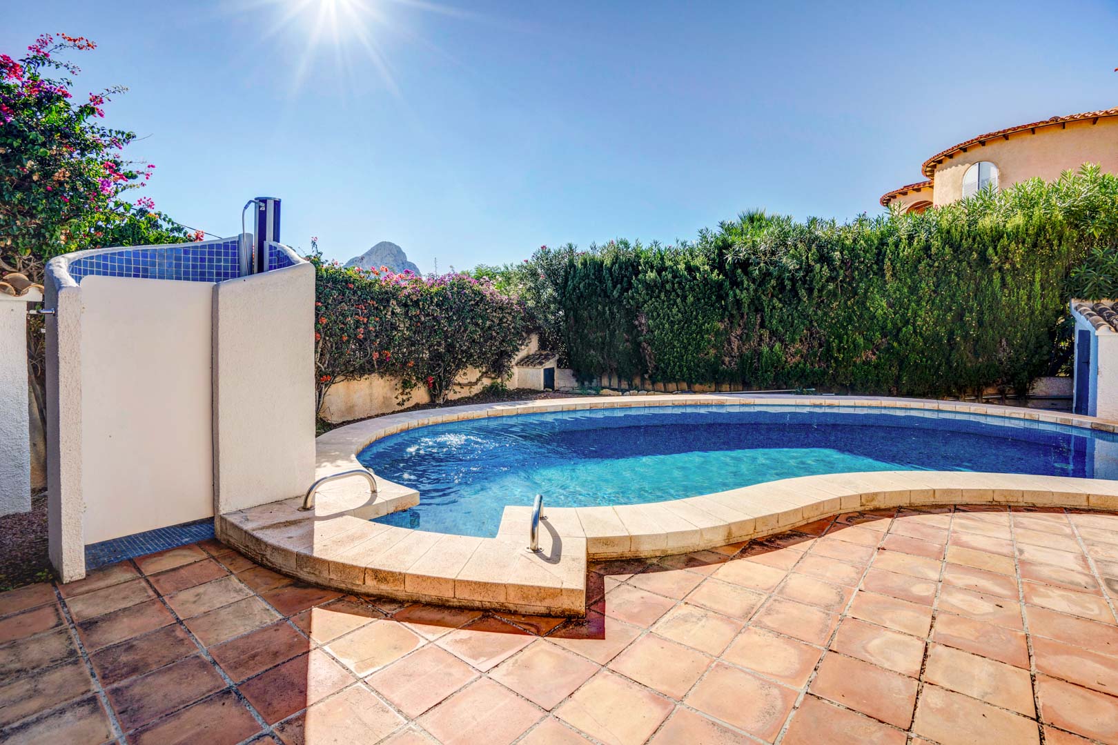 Piscine privée de forme incurvée avec terrasse en terre cuite, douche extérieure carrelée et vue sur le Peñón d'Ifach derrière des haies verdoyantes.