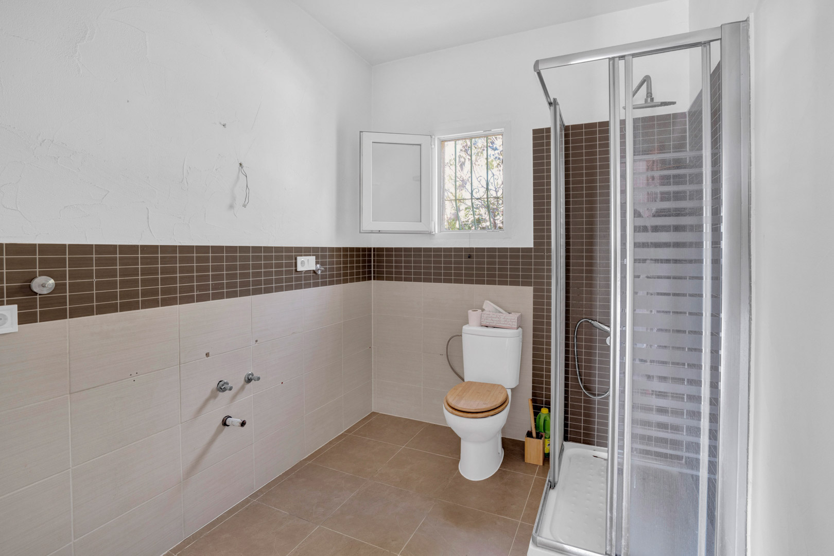 Salle de bain équipée d'une douche avec paroi vitrée, carrelage mosaïque marron et WC avec abattant en bois. Les murs sont revêtus de carreaux beiges et disposent d'une fenêtre.