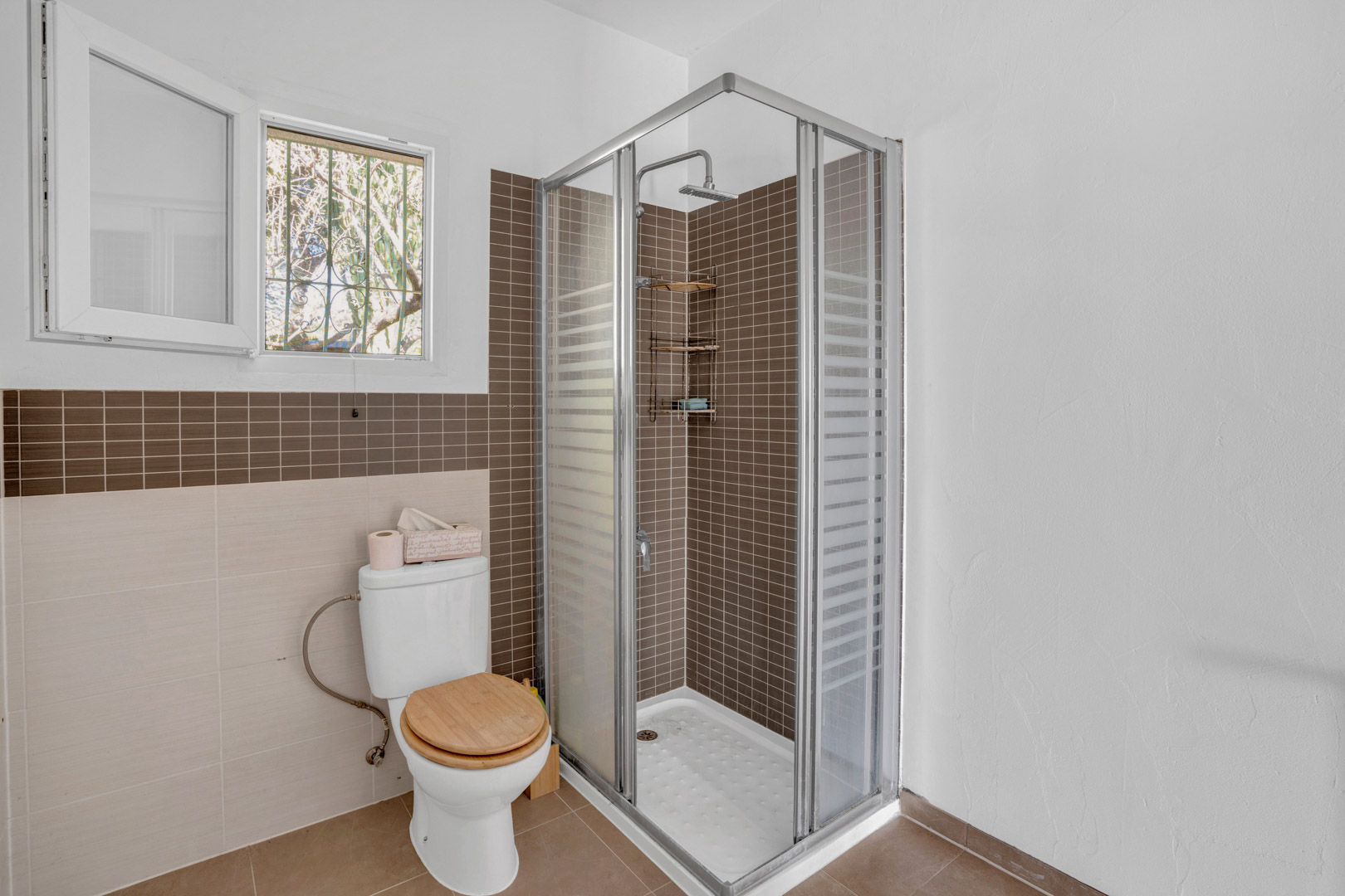 Salle de bain fonctionnelle avec douche d'angle en mosaïque marron, paroi vitrée, toilettes avec abattant en bois et fenêtre pour une aération naturelle.