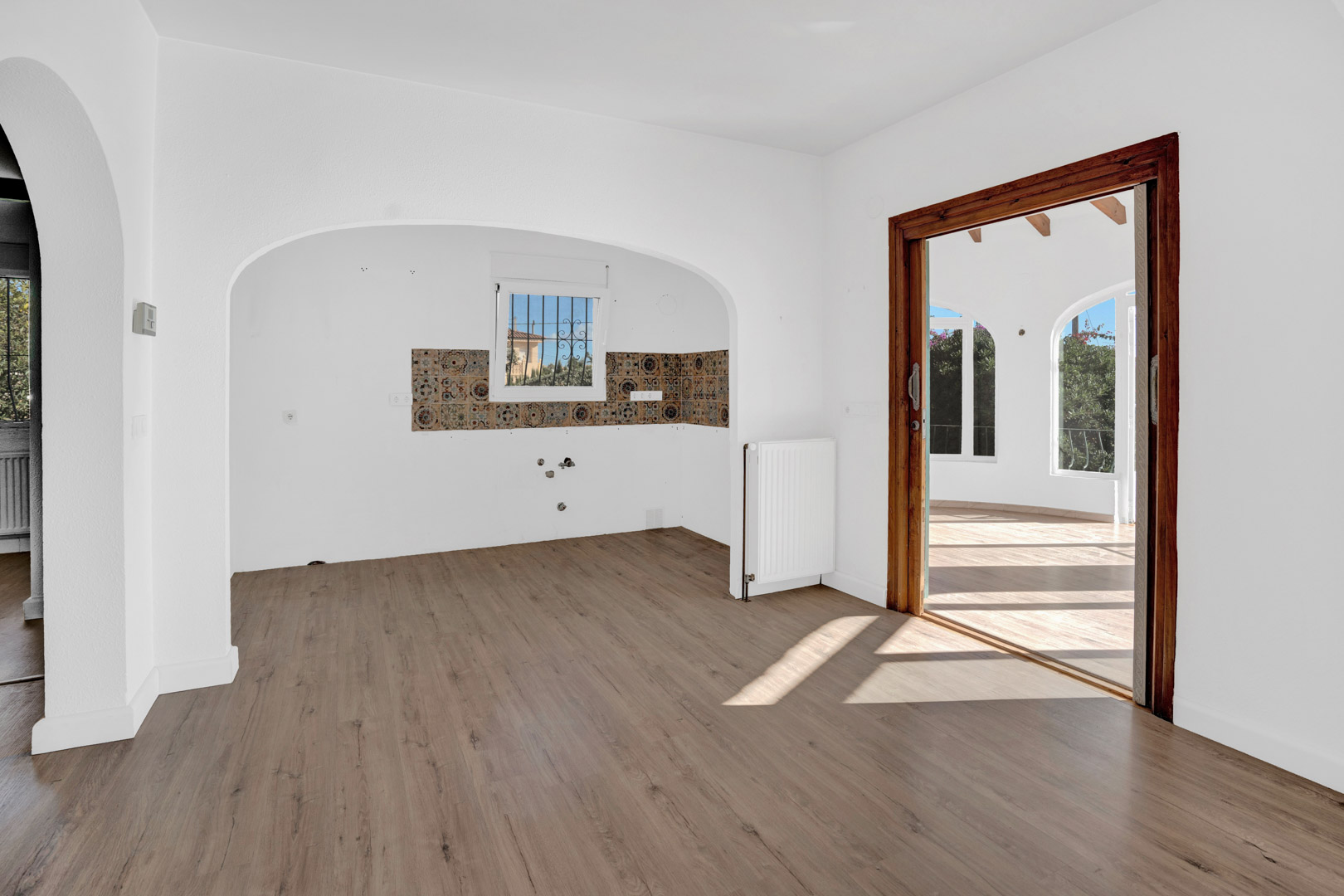 Salon avec sol stratifié, radiateur et alcôve de cuisine ornée de carreaux décoratifs. Une porte coulissante en bois mène à la terrasse vitrée lumineuse.