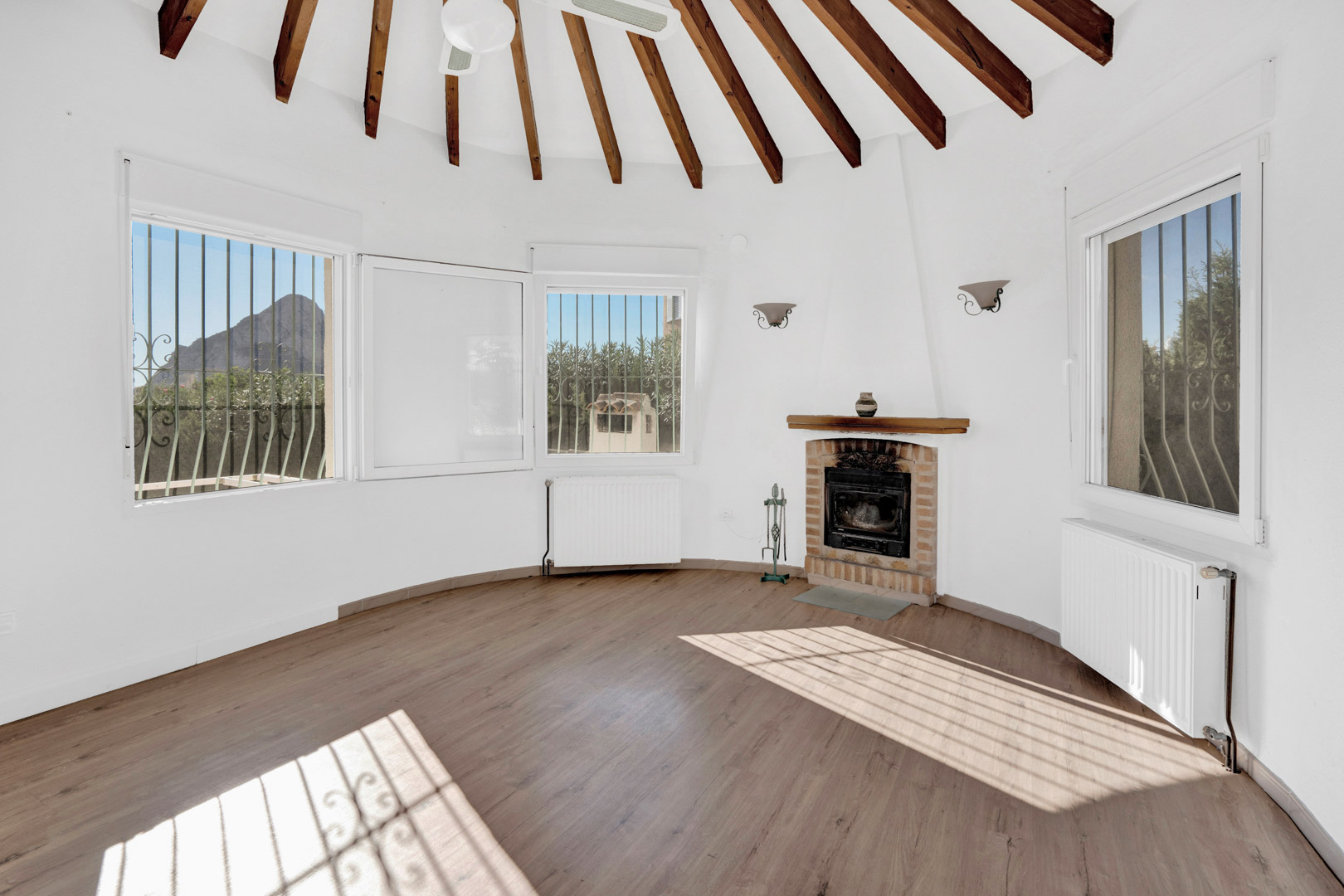 Salon circulaire avec plafond à poutres apparentes en éventail, cheminée traditionnelle en brique et fenêtres offrant une vue sur le Peñón de Ifach.