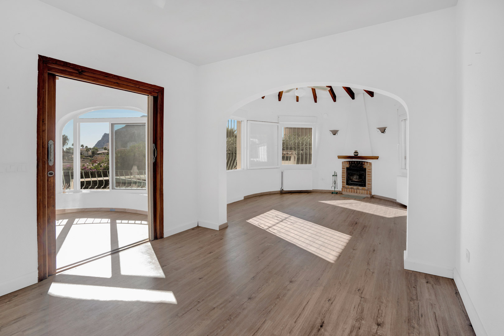 Salon lumineux avec cheminée en brique, poutres en bois apparentes et accès à une véranda offrant une vue dégagée sur le Peñón de Ifach.