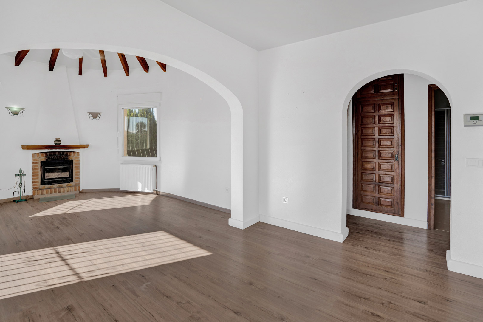 Salon lumineux avec cheminée en briques, poutres en bois apparentes et sol stratifié. Comprend des arches architecturales, un chauffage par radiateur et une porte en bois massif.