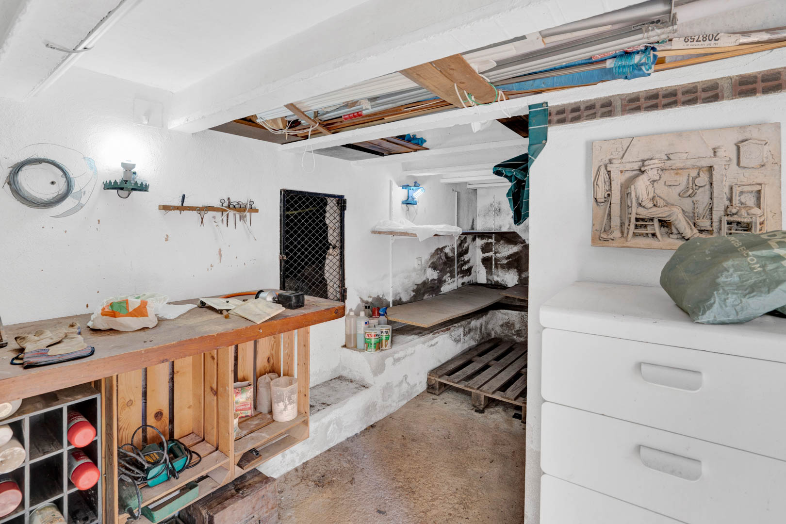 Sous-sol semi-enterré fonctionnel avec établi en bois, murs blanchis à la chaux et espaces de rangement polyvalents pour le bricolage ou le stockage.