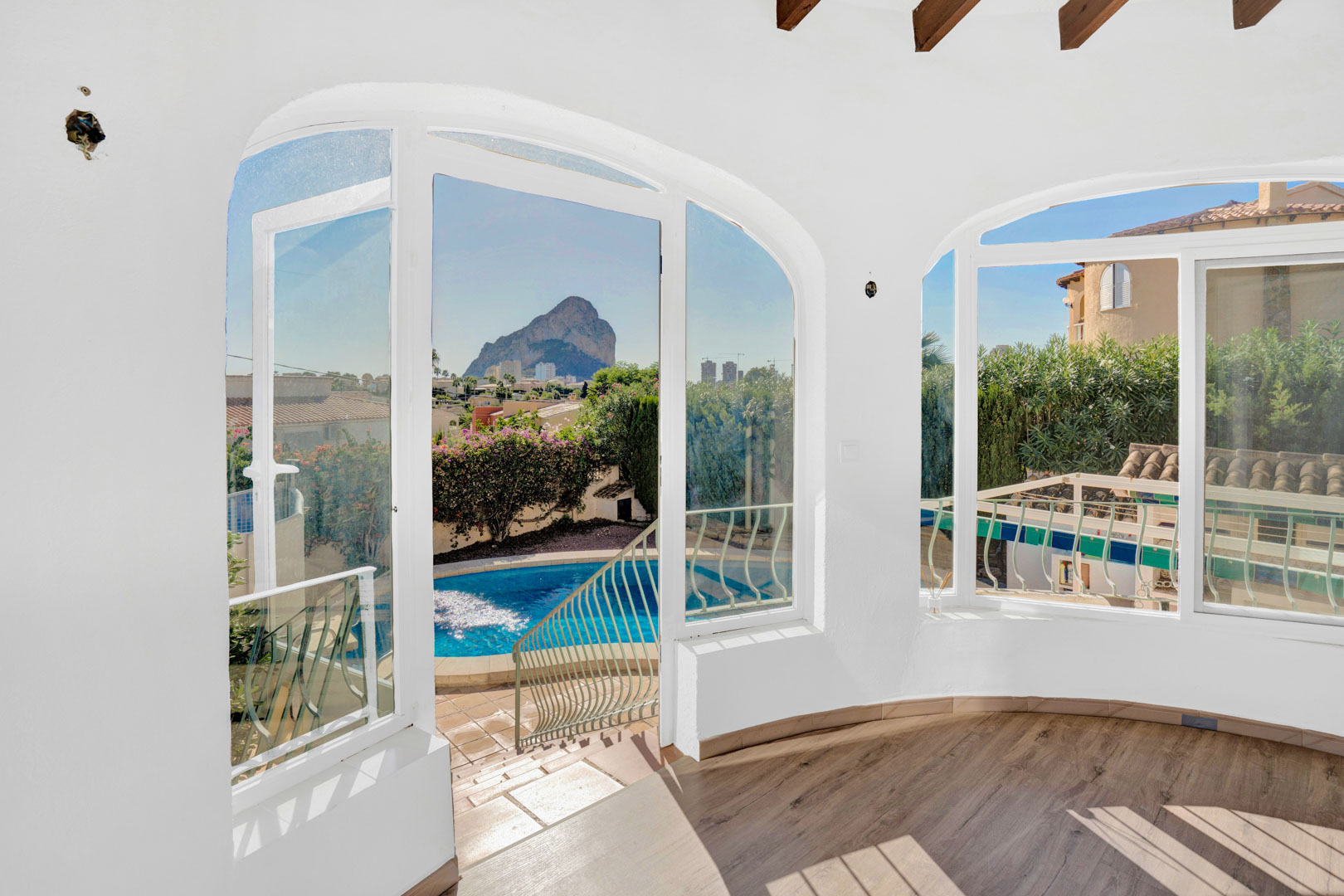Terrasse vitrée avec parquet, poutres apparentes et grandes fenêtres cintrées offrant une vue directe sur le Peñón de Ifach et l'espace piscine.