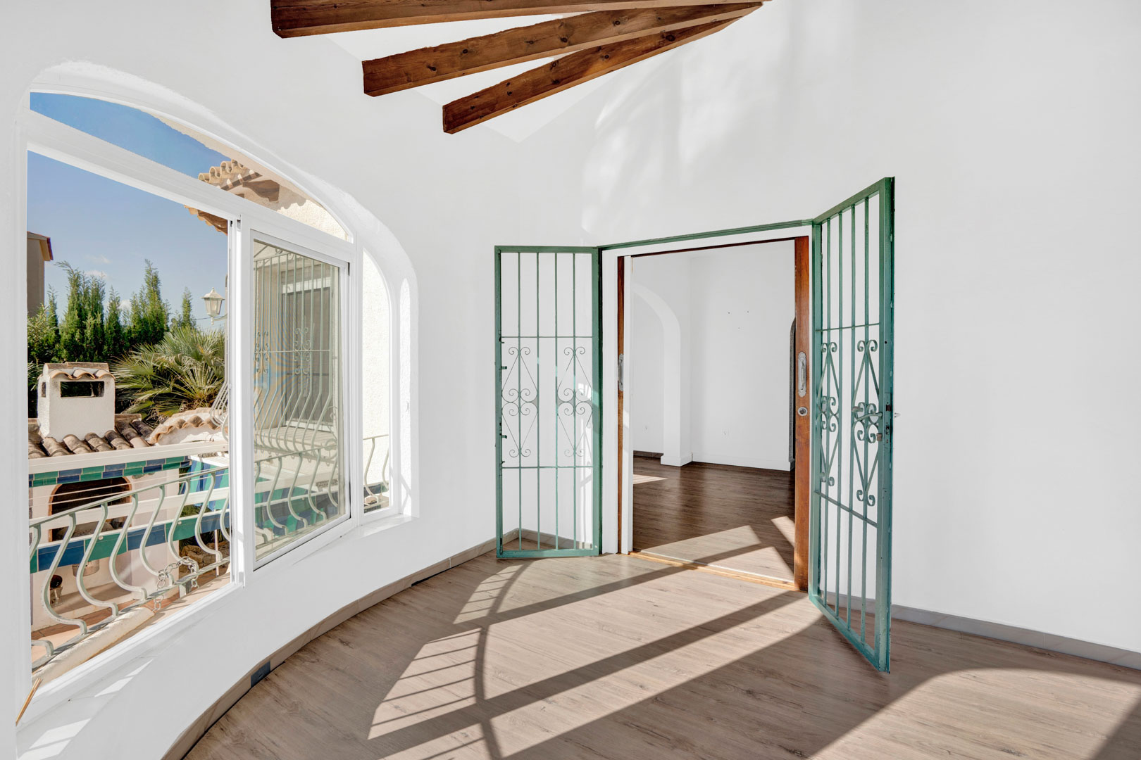 Terrasse vitrée avec poutres décoratives en bois, sol stratifié et fenêtres en arc offrant une vue dégagée et un accès sécurisé vers l'intérieur.