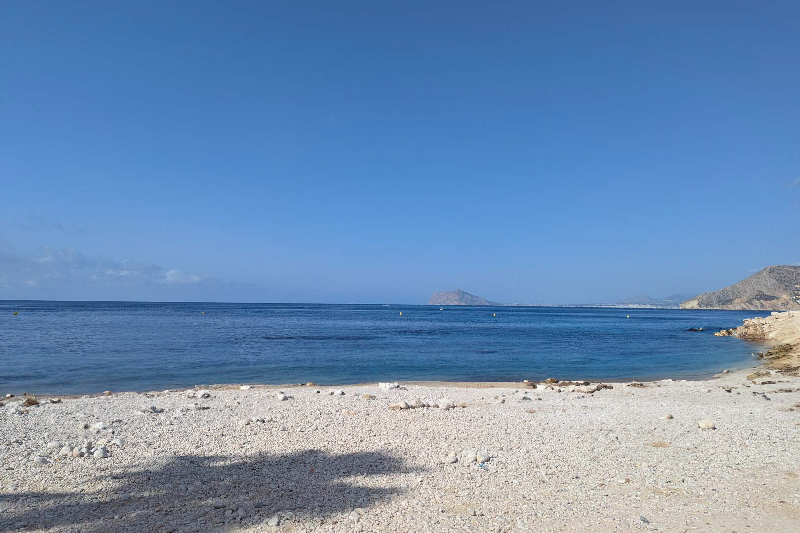 Vue d'une crique de galets blancs aux eaux calmes à Calpe, montrant le littoral méditerranéen et les montagnes au loin sous un ciel dégagé.