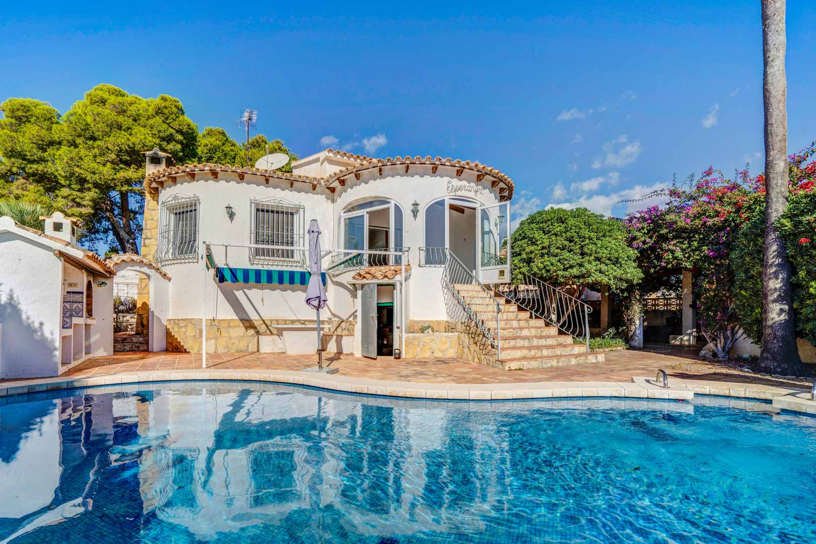 Vue extérieure de la villa avec piscine privée, façade blanche arrondie et escalier en pierre accédant à la terrasse vitrée et au jardin arboré.