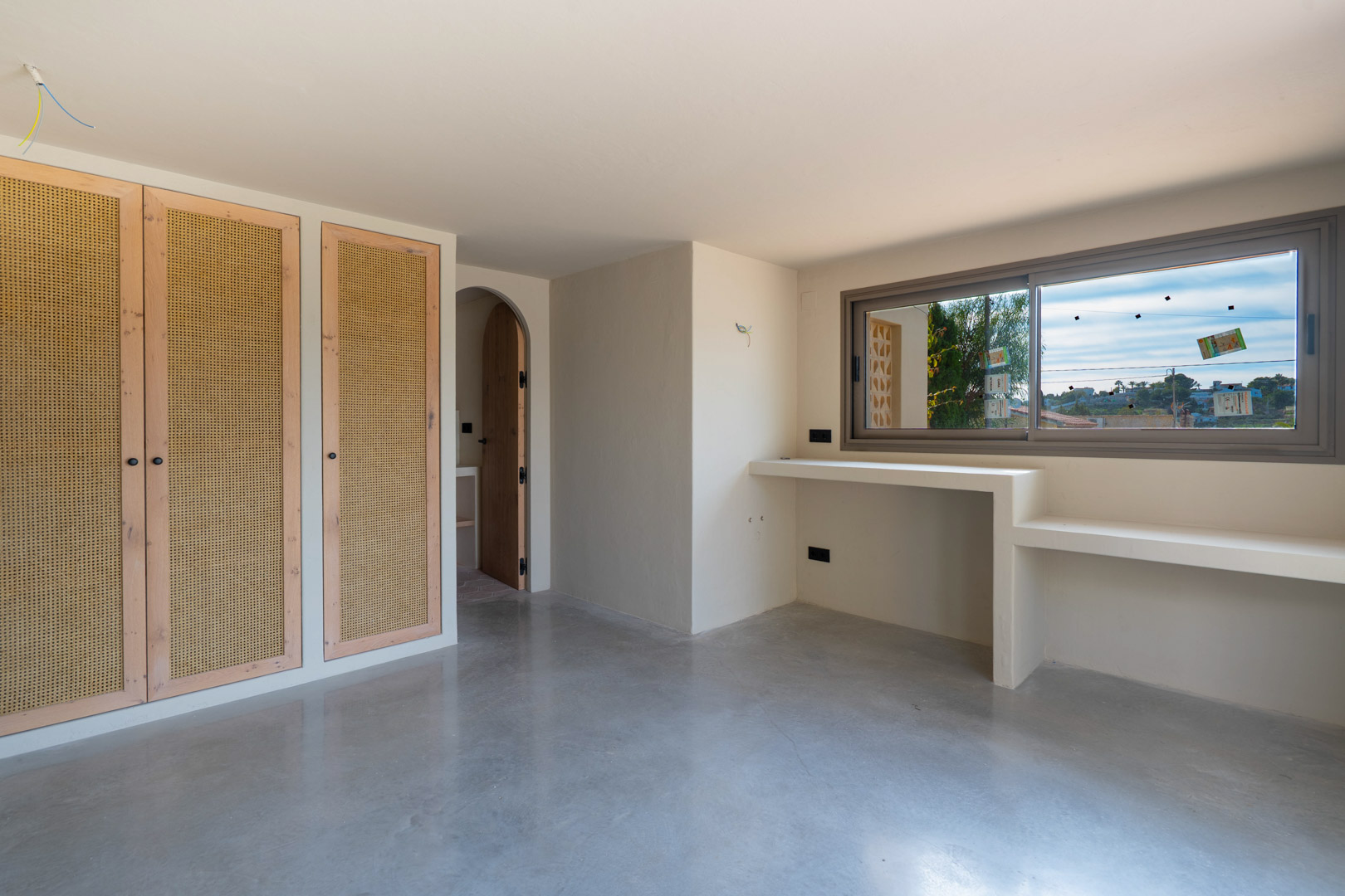 Chambre avec placards intégrés en bois et rotin, sol en béton poli et bureau maçonné sous la fenêtre. Une ouverture voûtée mène à la suite.