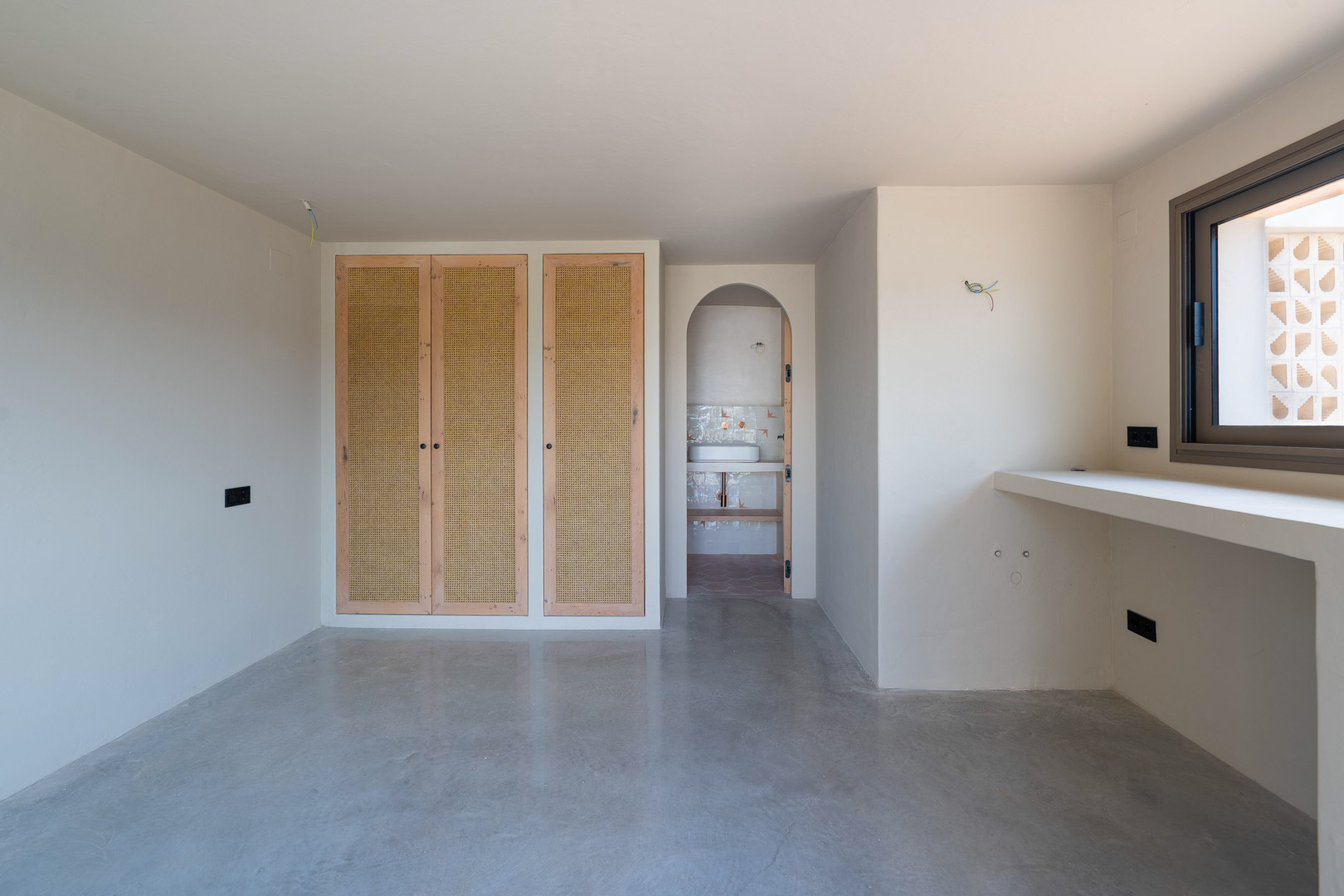 Chambre avec placards intégrés en chêne et rotin, sol en béton poli et ouverture en arche vers une salle de bain attenante aux finitions naturelles.