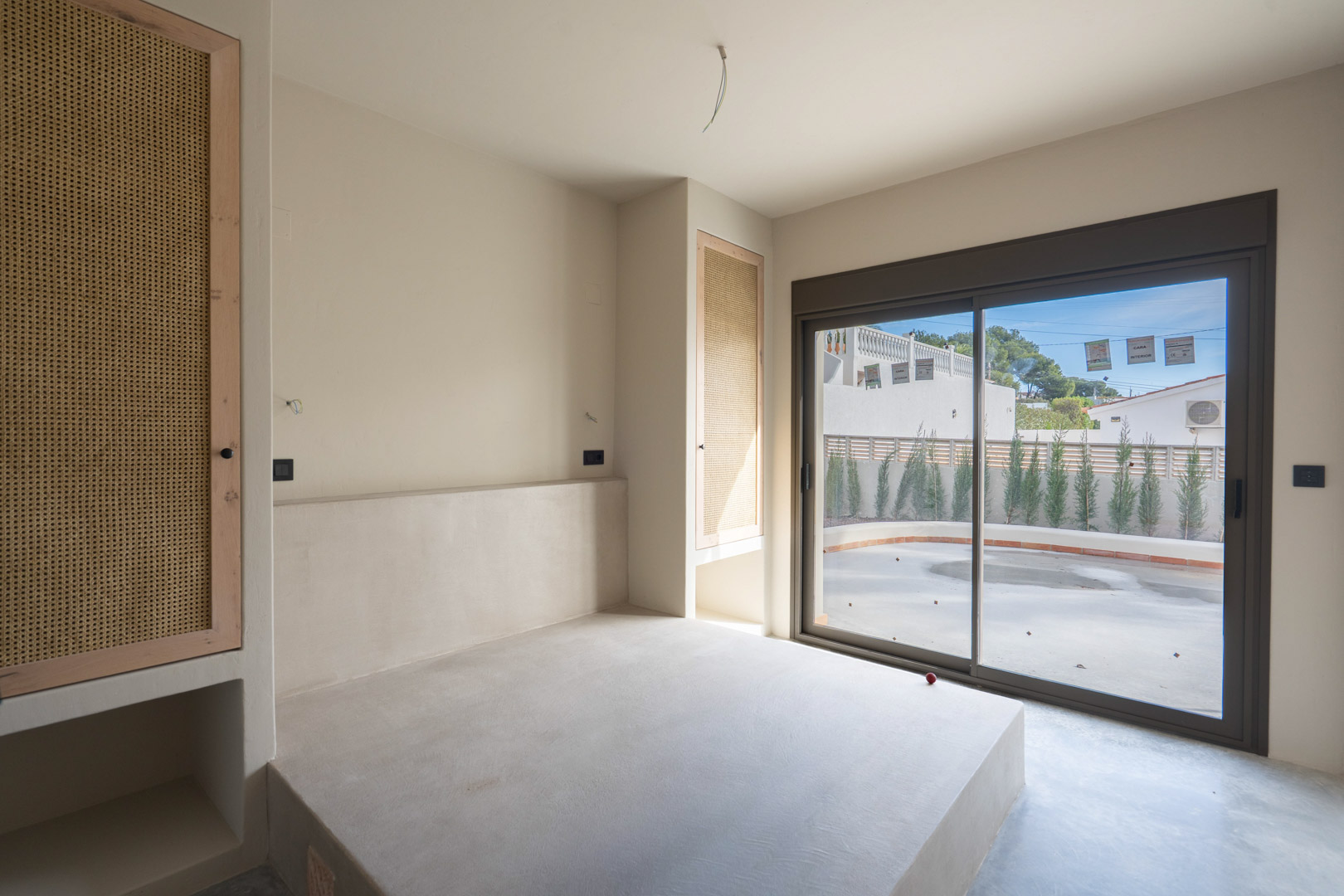 Chambre avec plateforme de lit en béton poli, armoires encastrées en chêne et rotin, et grande baie vitrée coulissante ouvrant sur la terrasse extérieure.