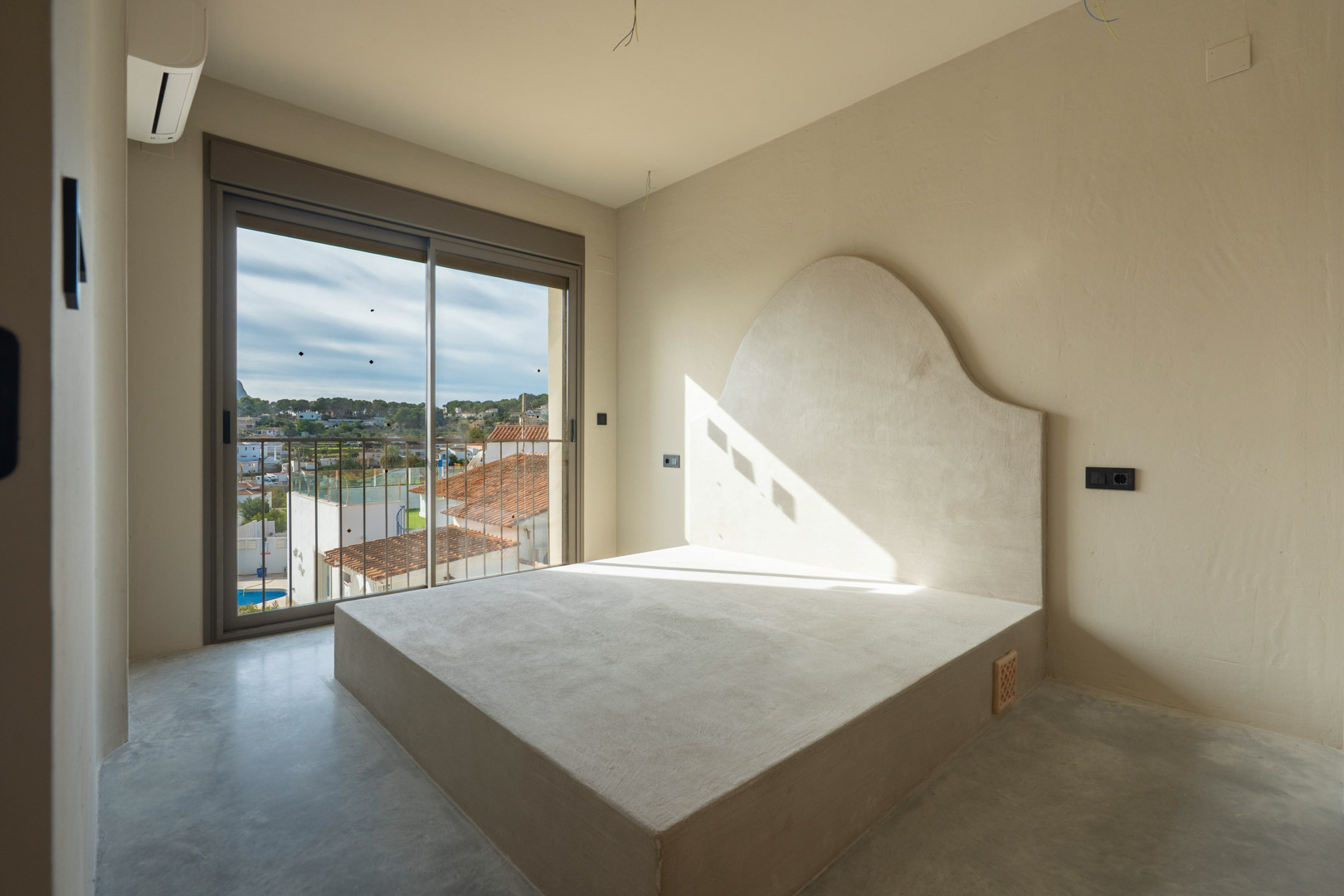 Chambre avec sommier intégré et tête de lit incurvée en argile, sol en béton poli et accès direct à un balcon offrant une vue sur les environs.