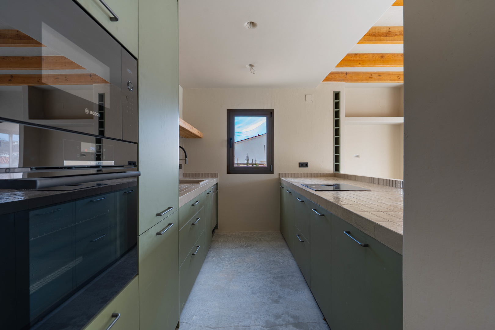 Cuisine design avec meubles vert olive, appareils Bosch intégrés et plans de travail carrelés. Elle dispose de poutres en bois apparentes et d'un sol en béton poli.