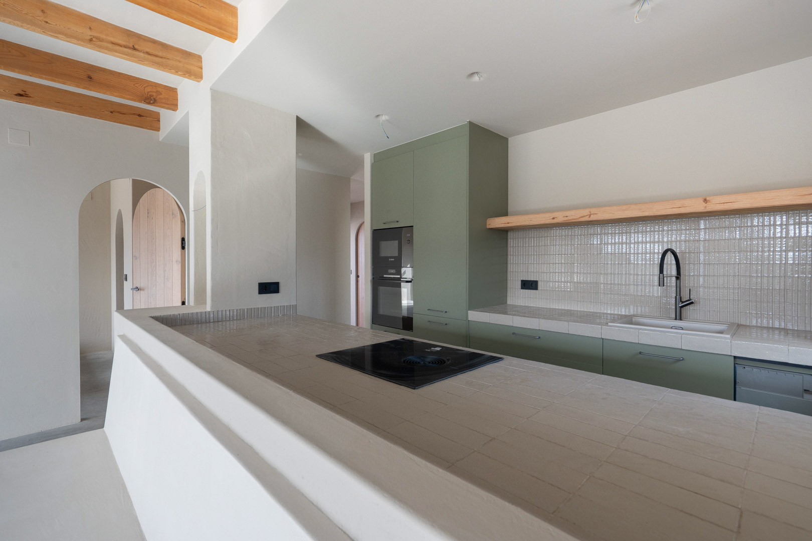 Cuisine design avec meubles vert sauge, plans de travail en porcelaine Ragno et plaque Bora. Présente des poutres en bois apparentes et des finitions en argile.