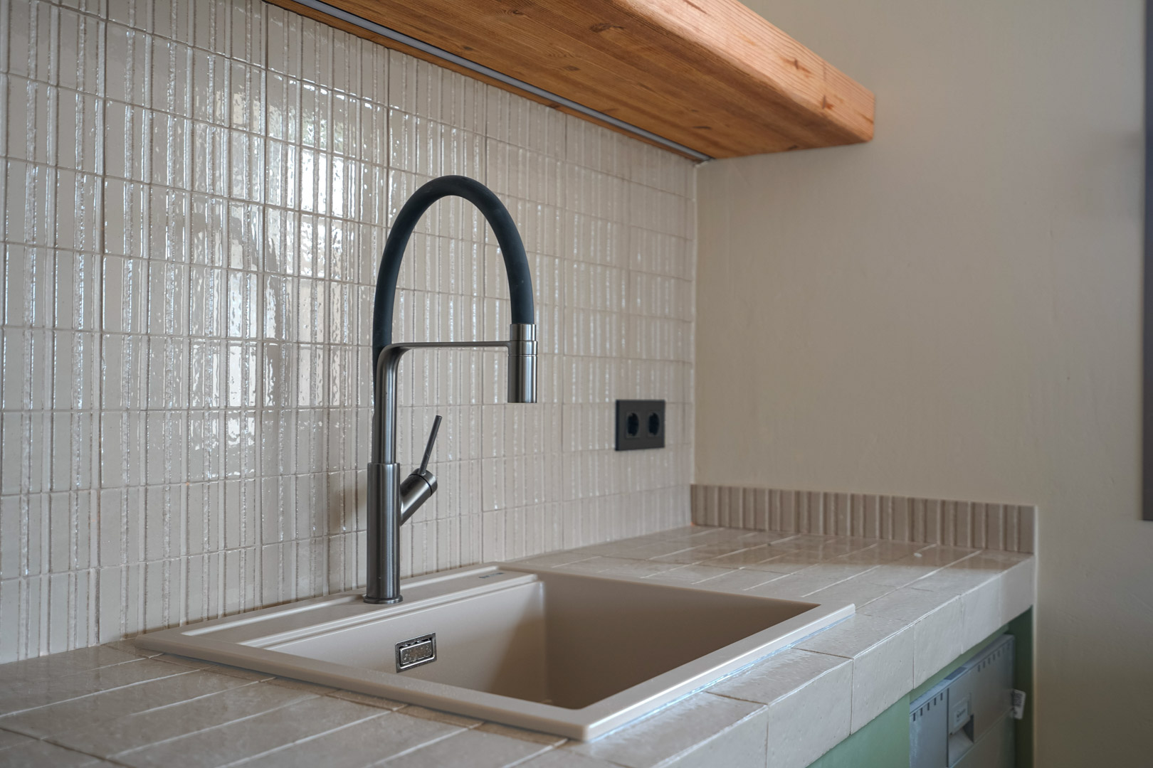 Détail de la cuisine avec plan de travail en grès cérame et évier intégré. Robinetterie moderne à col flexible sur une crédence en mosaïque verticale et étagère en bois massif.