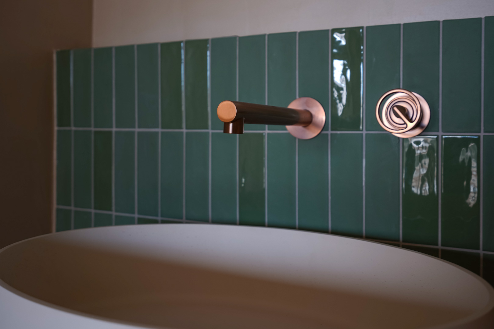 Détail de salle de bain avec robinetterie murale en cuivre, carrelage vertical vert émeraude brillant et vasque à poser blanche mate.
