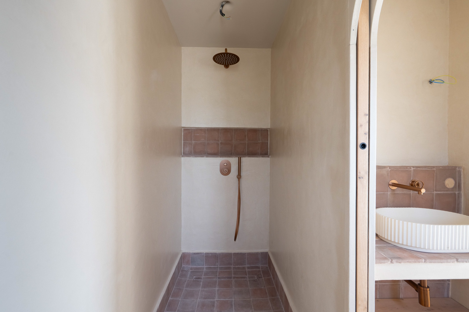 Douche à l'italienne avec finitions en argile naturelle, robinetterie en bronze et sol en terre cuite. Vue sur le lavabo vasque cannelé et sa robinetterie murale.