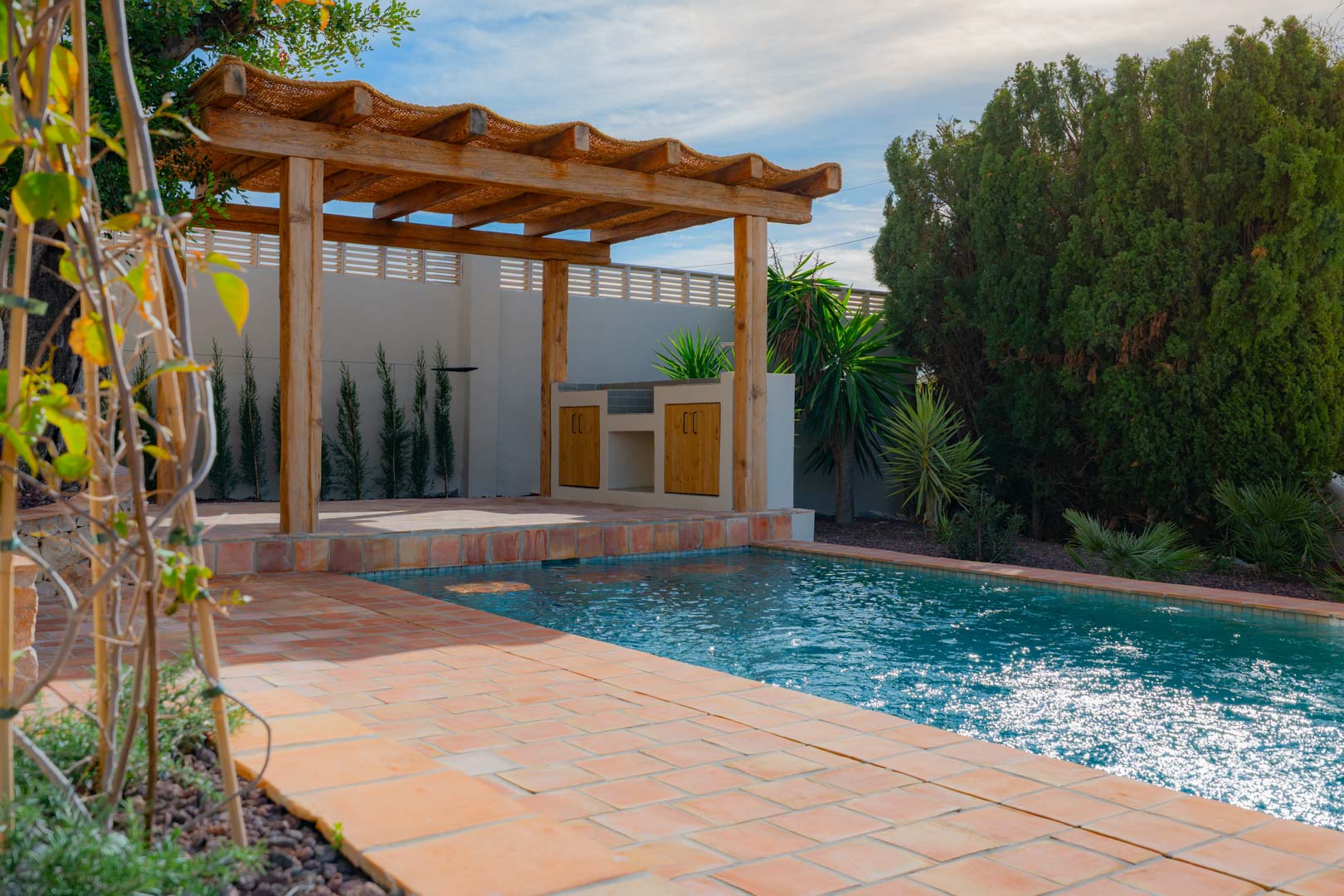 Espace extérieur avec piscine en mosaïque bleue, terrasse en terre cuite et pergola en bois abritant une cuisine d'été, le tout entouré d'un jardin méditerranéen.
