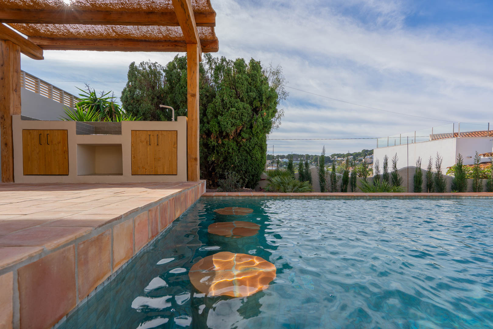 Espace piscine avec sièges immergés, cuisine d'été sous pergola en bois et revêtement de sol en terre cuite méditerranéenne.