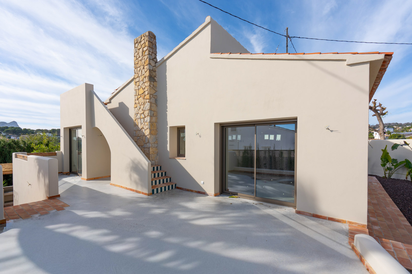 Extérieur de la villa avec terrasse en béton poli, cheminée en pierre naturelle et escalier orné de carreaux. Vue sur le Peñón de Ifach à l'horizon.
