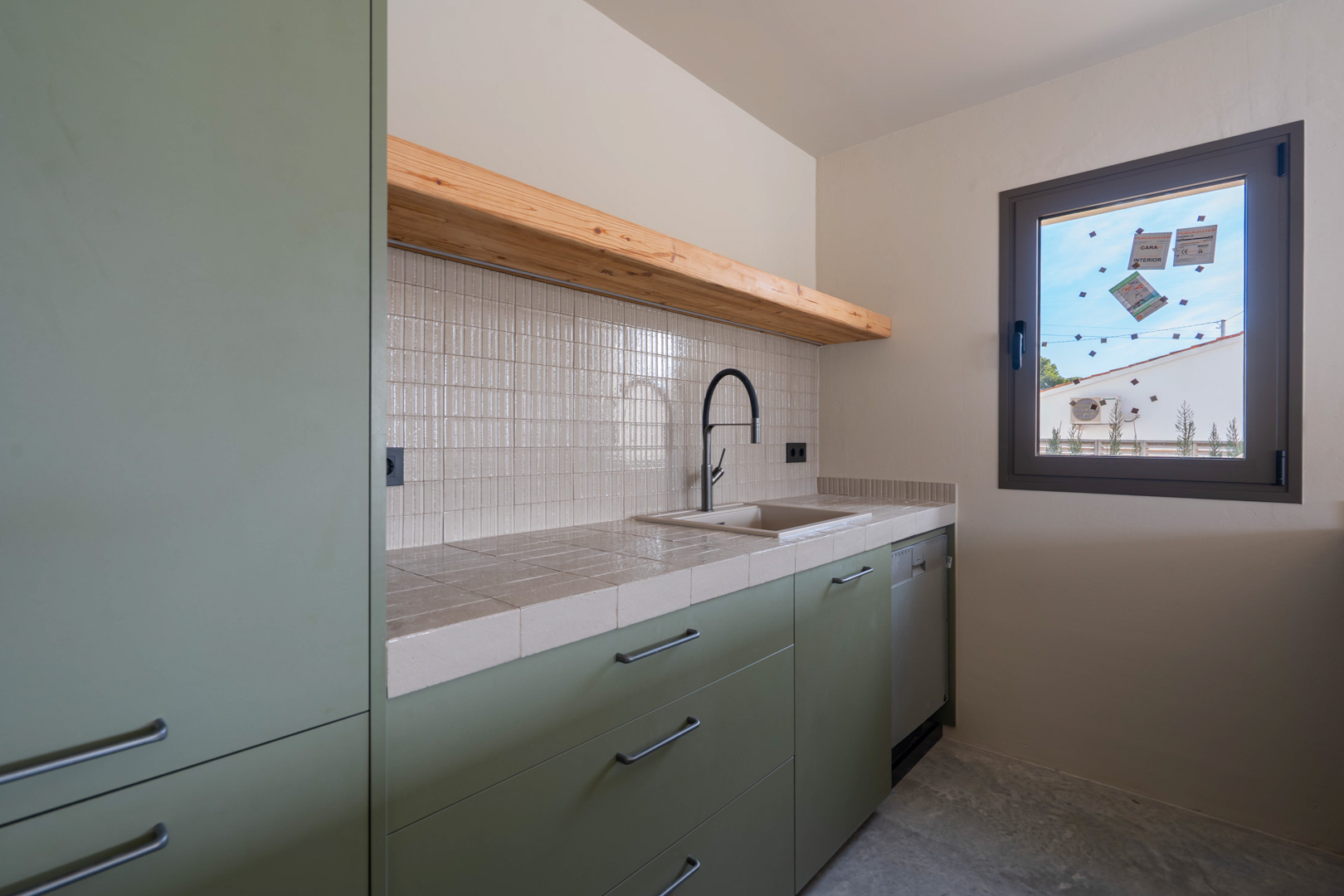 Kitchenette d'invités avec meubles vert sauge, plan de travail carrelé, étagère en bois massif et robinetterie noire sur sol en béton poli.