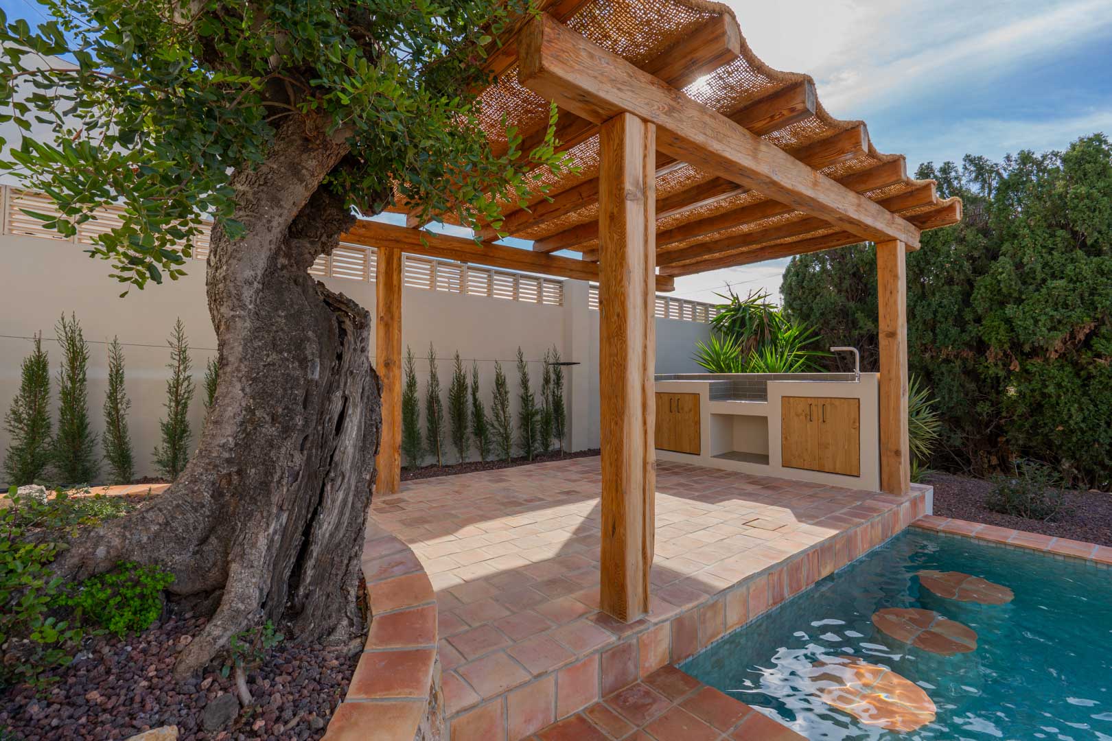 Pergola en bois avec toit en fibres naturelles sur sol en terre cuite, incluant une cuisine d'été et une piscine avec sièges immergés près d'un olivier ancien.