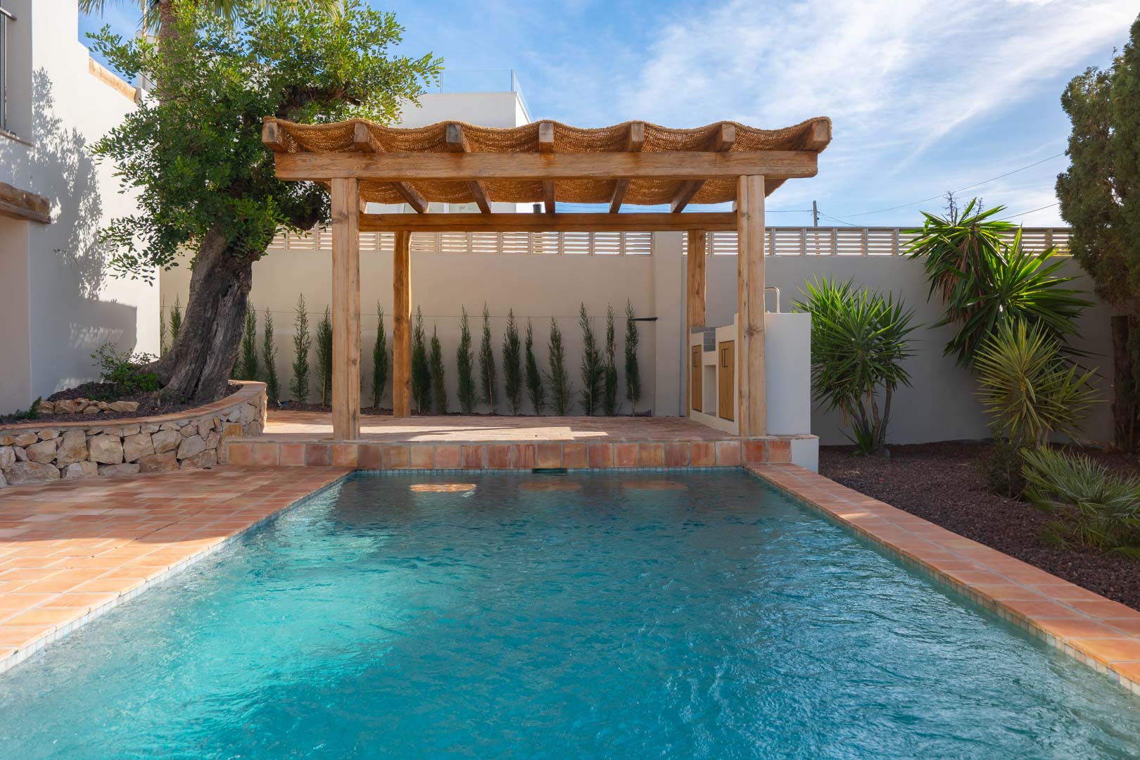 Piscine extérieure bordée de terre cuite avec une pergola en bois et un espace barbecue, le tout entouré d'un jardin méditerranéen aux essences variées.