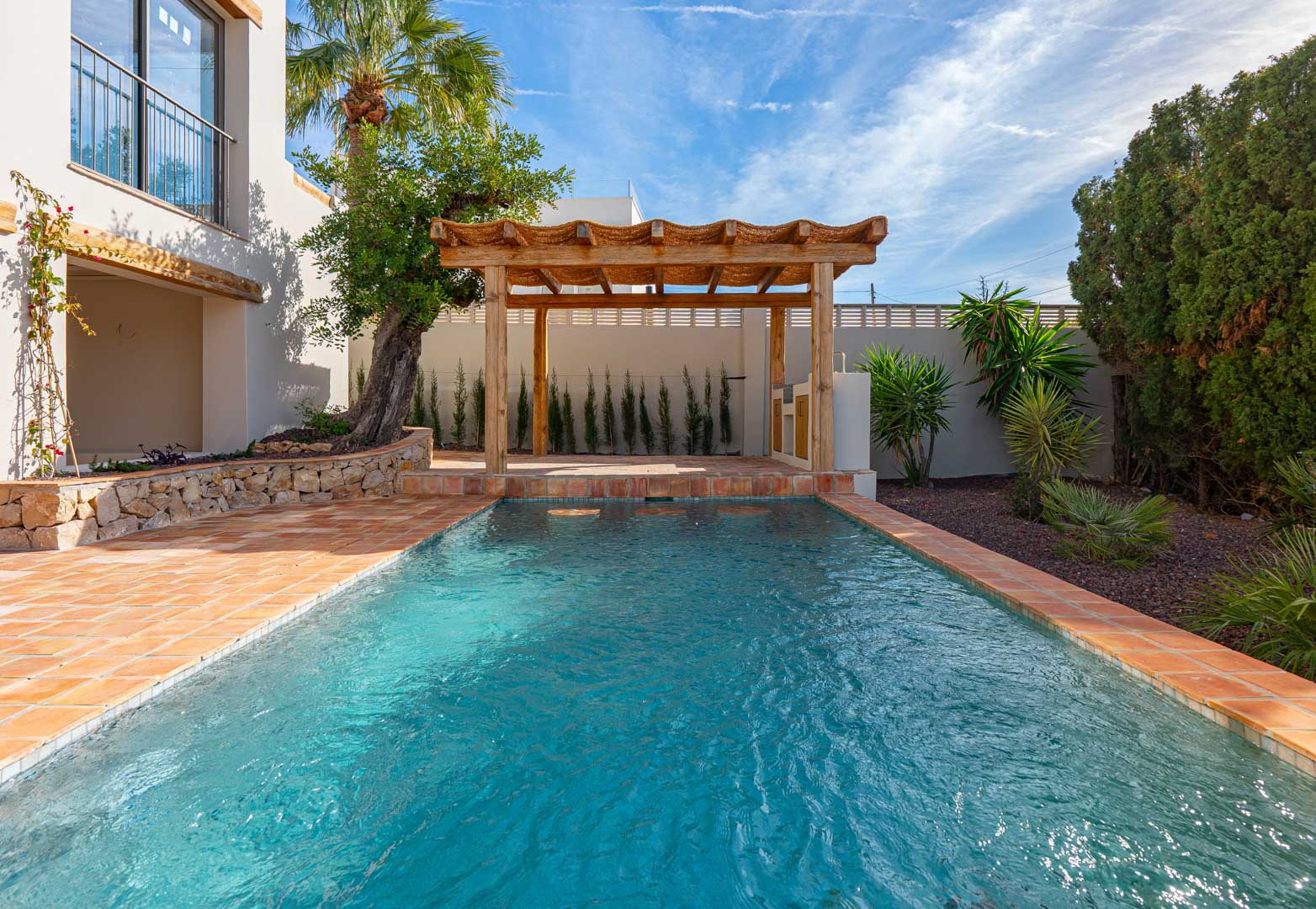Piscine rectangulaire bordée de terre cuite, avec pergola en bois et jardin méditerranéen paysager comprenant des murs en pierre et une végétation variée.