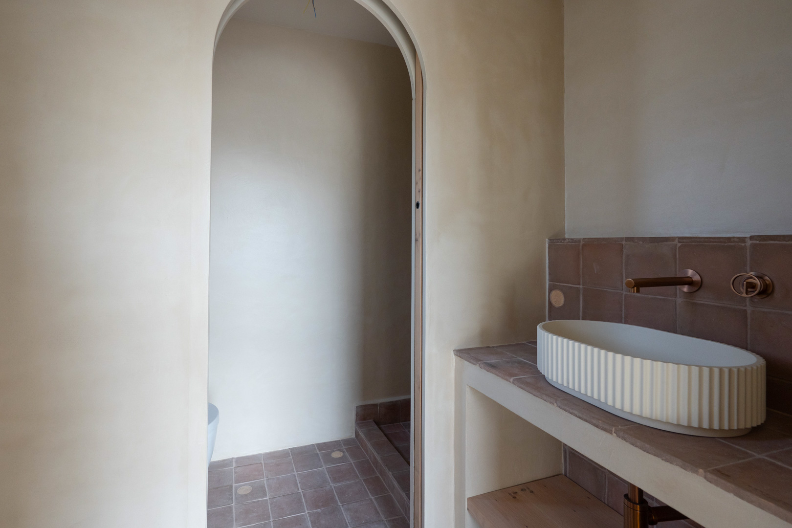 Salle de bain avec murs en argile, vasque cannelée et robinetterie encastrée en bronze. L'espace se distingue par son arche élégante et ses carreaux en terre cuite.