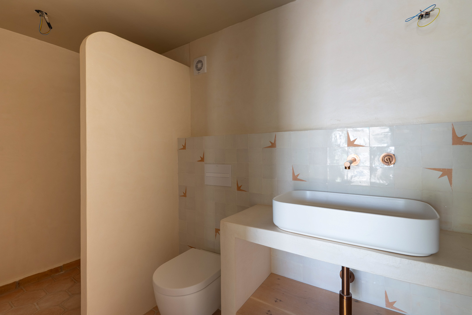 Salle de bains avec vasque à poser sur plan maçonné, robinetterie encastrée en cuivre et finitions en argile. Sol en terre cuite et WC suspendu au design épuré.