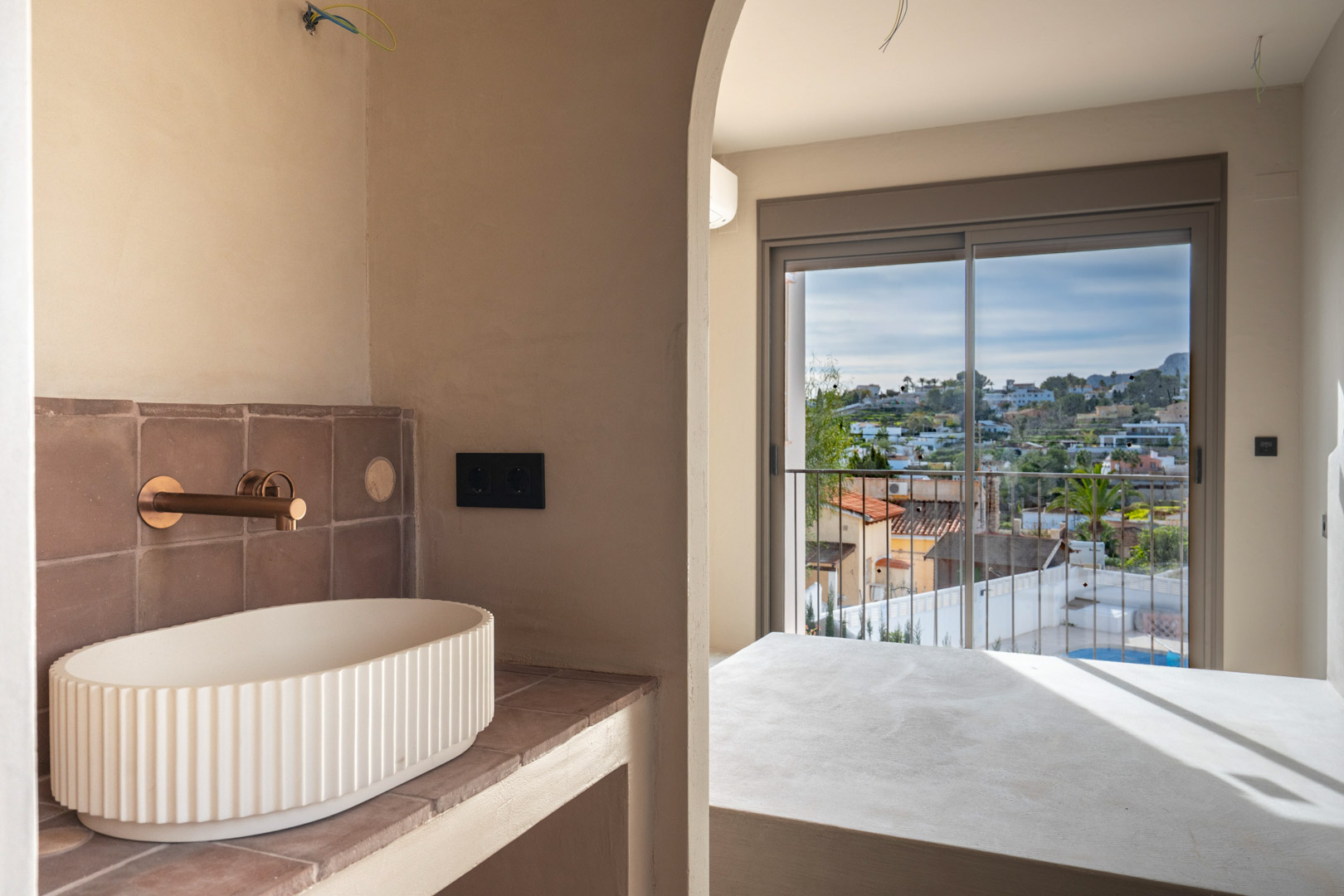 Salle de bains avec vasque cannelée et robinetterie en cuivre, offrant une vue à travers une arche vers la chambre et son balcon sur l'extérieur.